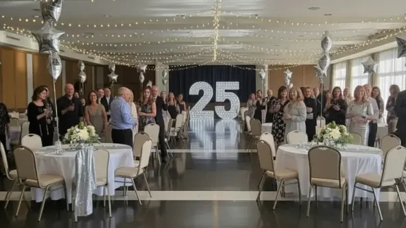 A group of people standing for a photo in Murray Hall. they are celebrating a 25th wedding anniversary. The party is silver themed with a big silver 25 in the center of the hall.