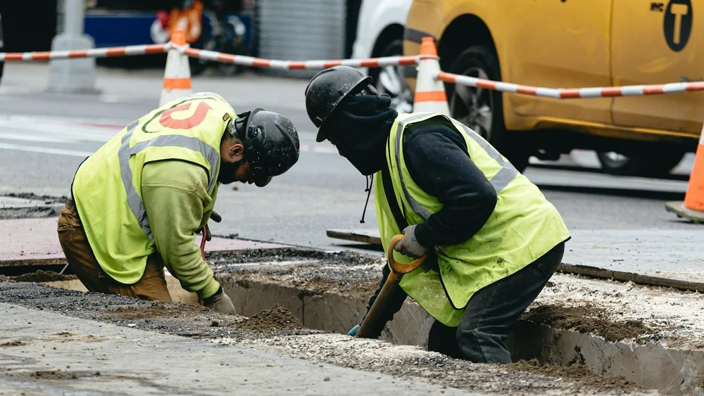 Zwei Arbeiter in Warnwesten heben einen schmalen Graben in einer Straße aus