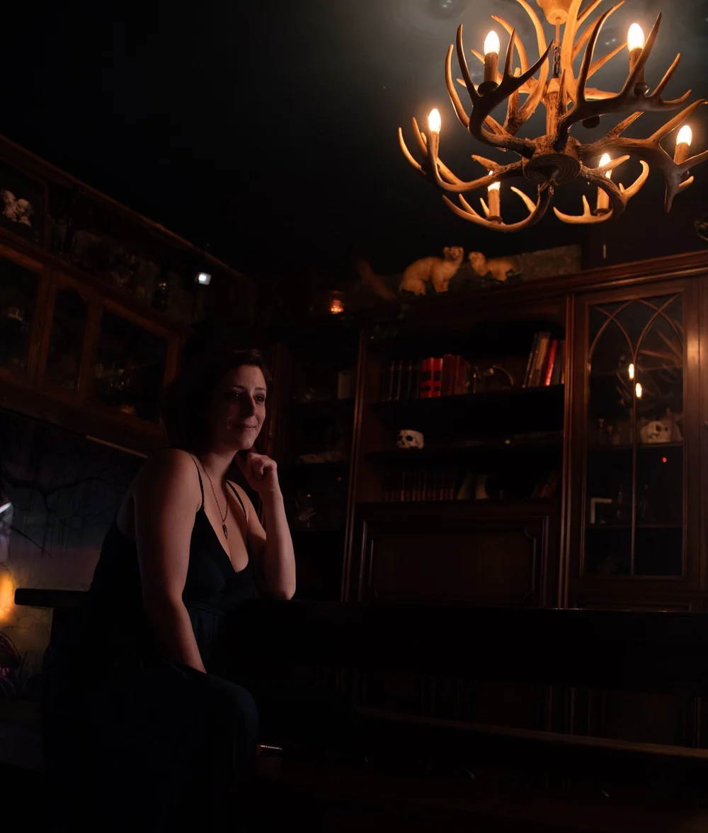 Rebecca assise sous le lustre des Trois Corbeaux, devant la bibliothèque et les vitrines en bois.