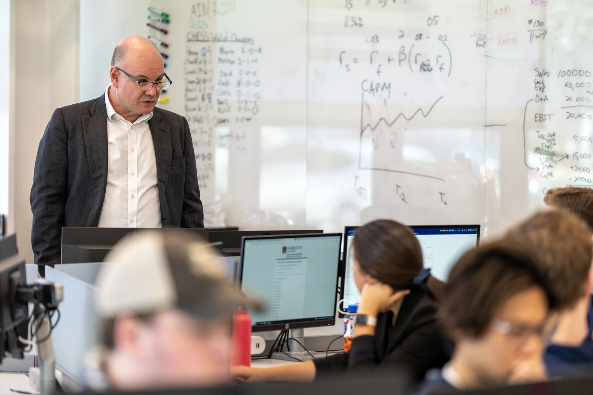 Joseph Wall teaching in a classroom, with students at computers and financial concepts on the whiteboard.