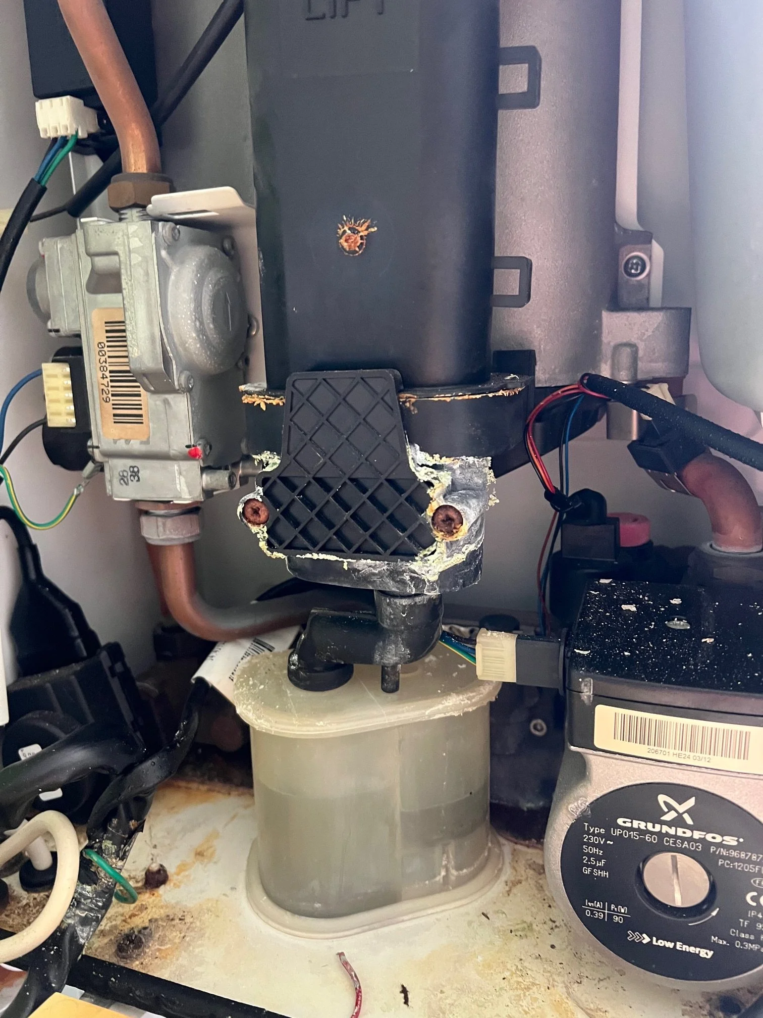 Close-up of a cracked sump on an Ideal Logic boiler showing distorted black plastic, corroded screw heads, and yellowed mineral deposits from long-term condensate leakage
