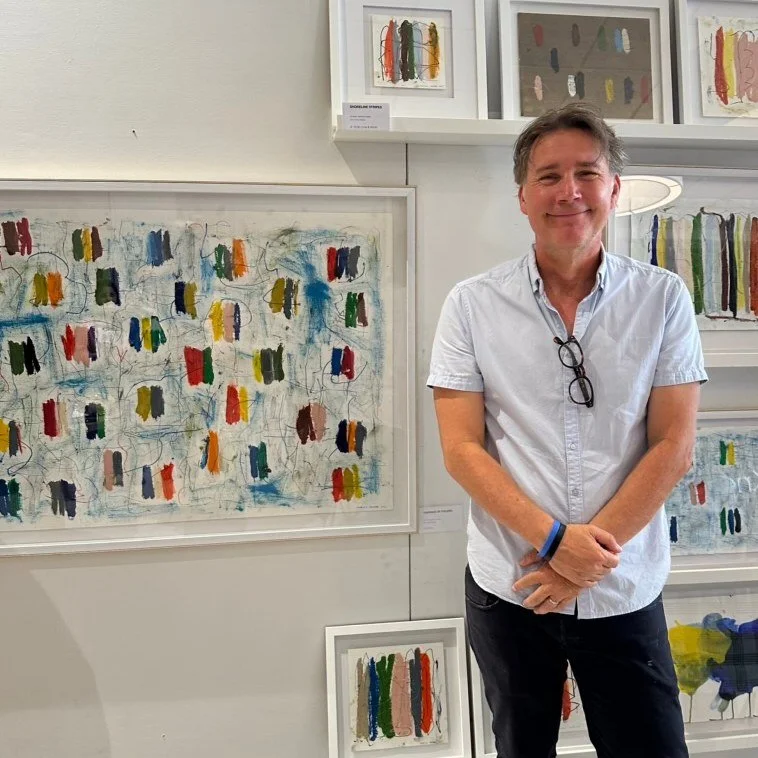Artist Marcus Bacher standing beside his framed abstract works at a gallery exhibition.