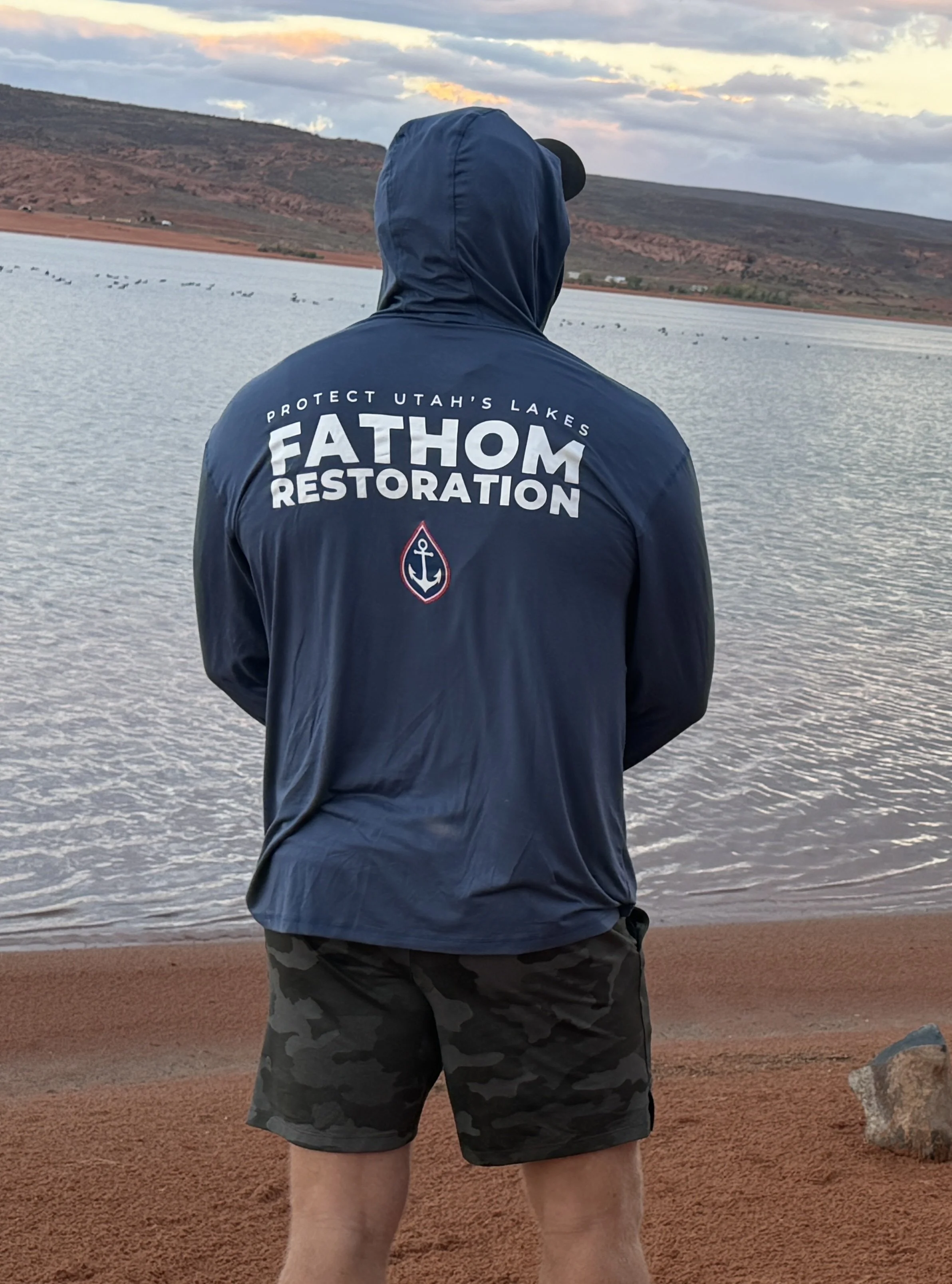A volunteer on the red sand shore at Sand Hollow in a Fathom Restoration hoodie that reads Protect Utah's Lakes, facing the water