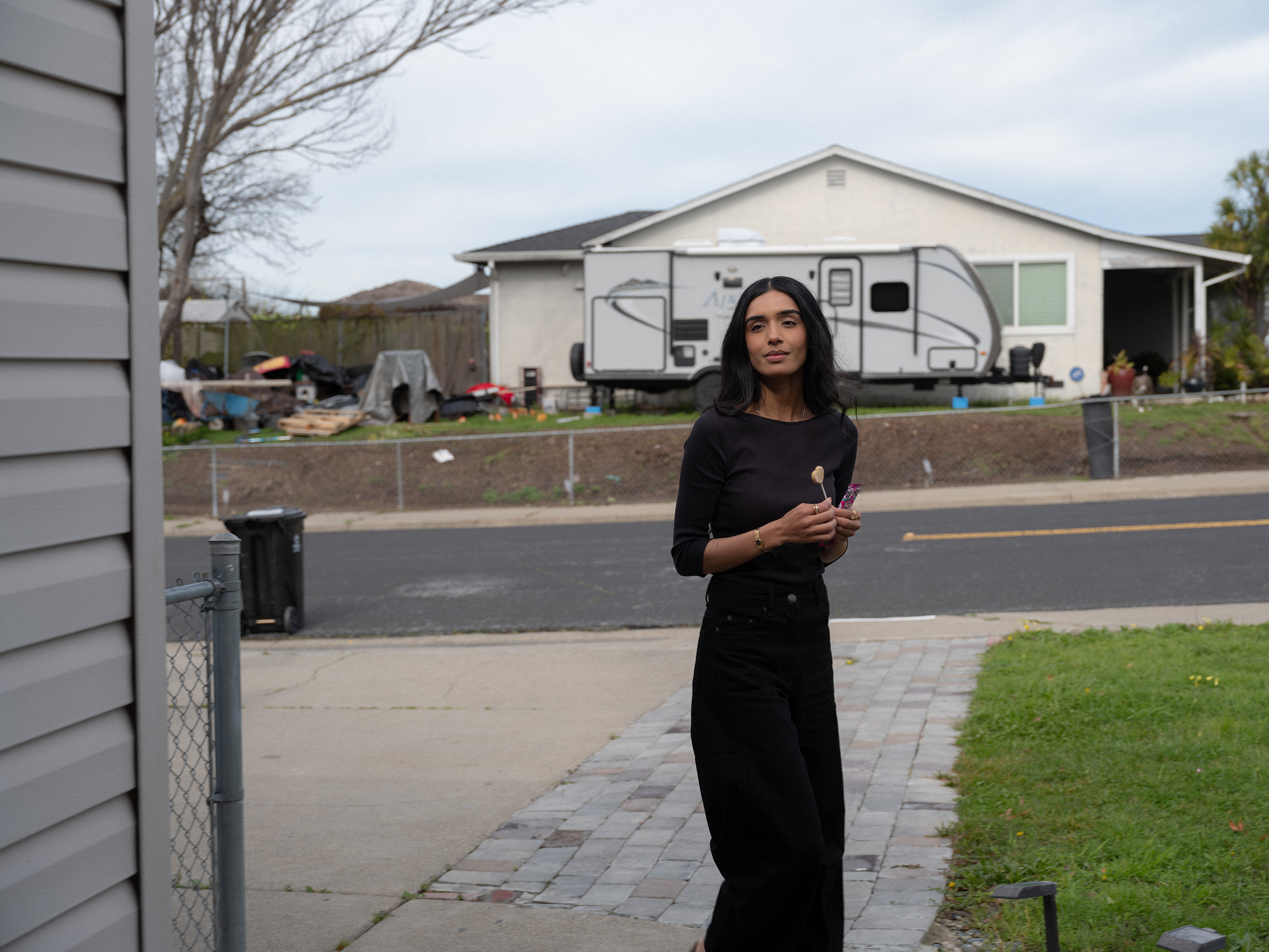 Sumbal stands on the pavement outside her California home, dressed in black, looking calmly toward the camera.
