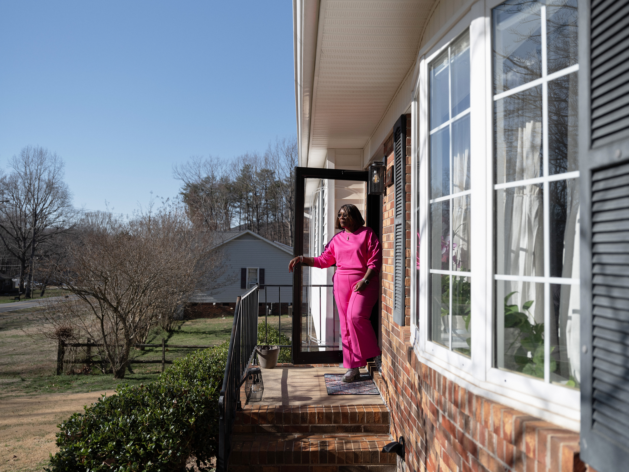 Jacqueline stands in the doorway of her North Carolina home, dressed in bright pink, on a sunny porch.