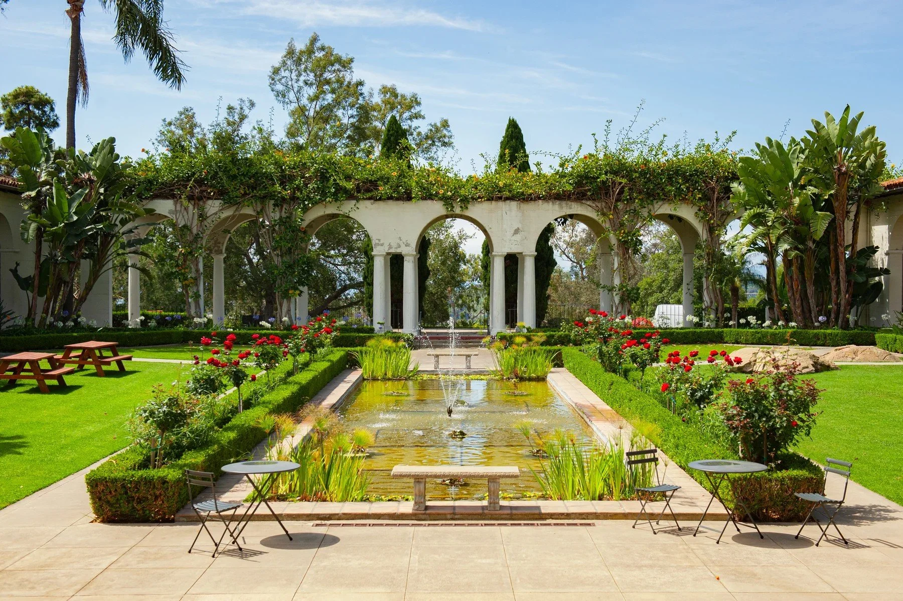 Long reflecting pool framed by clipped hedges, palms, and white arches at Riviera Park