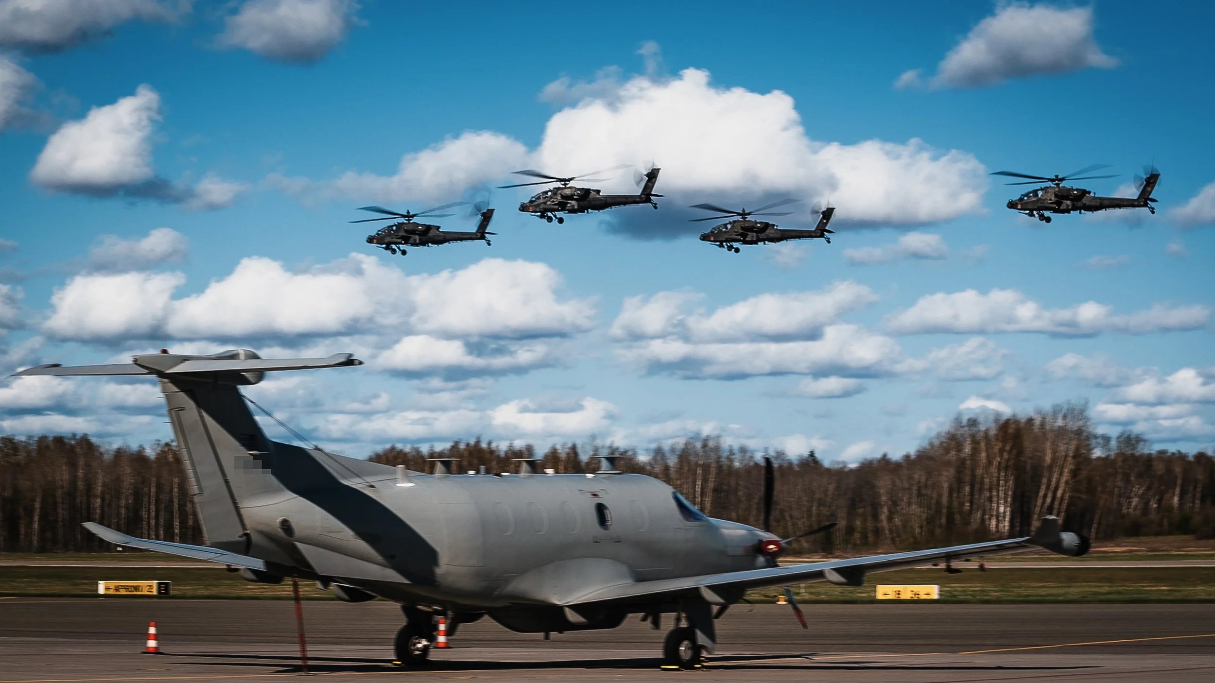 Four Apache helicopters in formation passing over a fixed-wing aircraft on tarmac