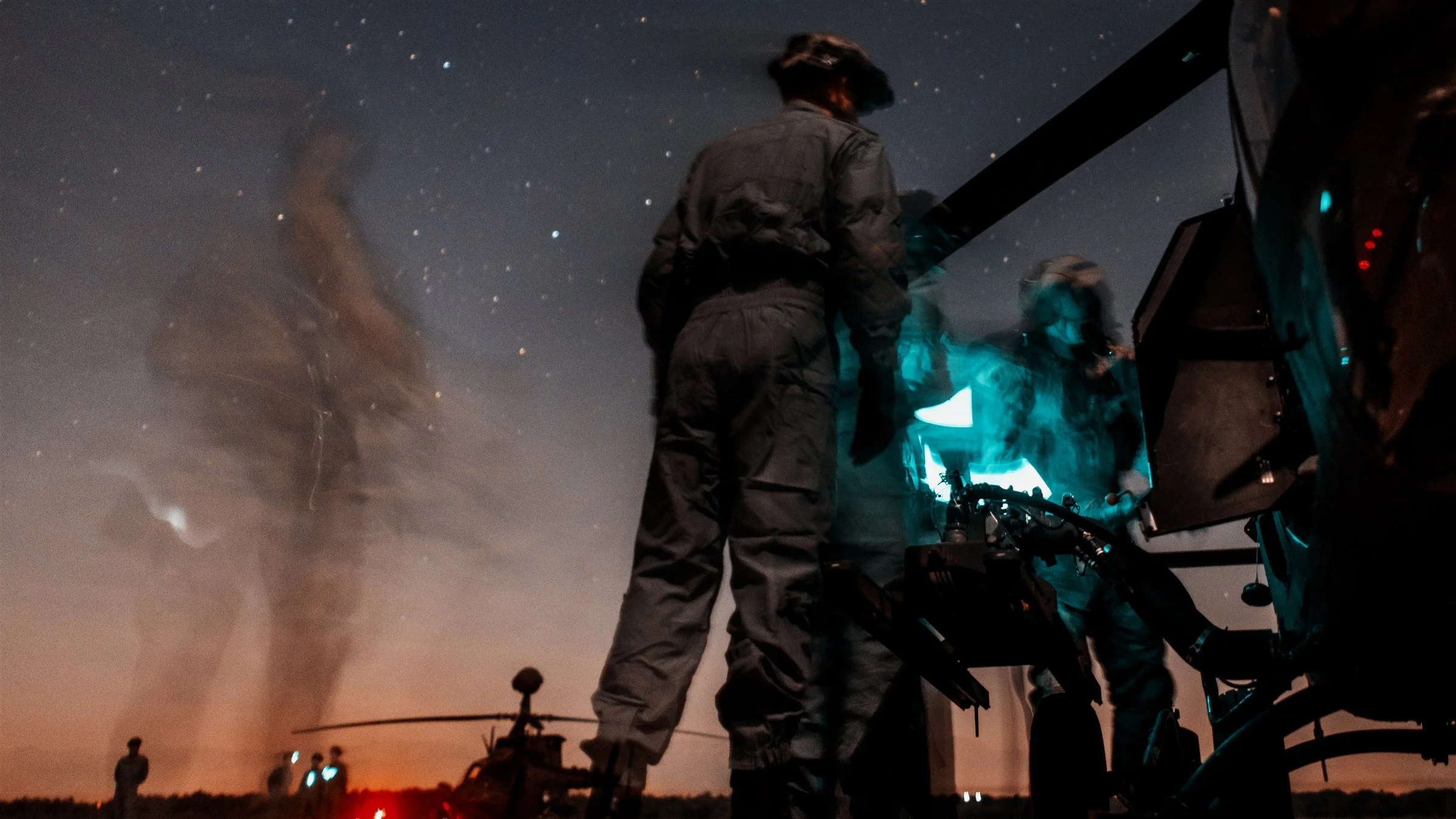 Helicopter crew member silhouetted against twilight sky during night operations, motion-blur figures in background
