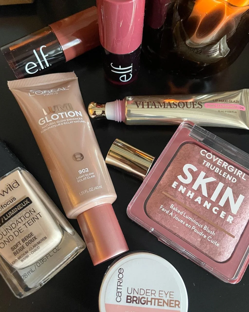 Flat lay of budget beauty dupes: e.l.f., L'Oreal Lumi Glotion, Vitamasques Lip Maxi Glaze, COVERGIRL TruBlend blush, Catrice Under Eye Brightener, and wet n wild foundation