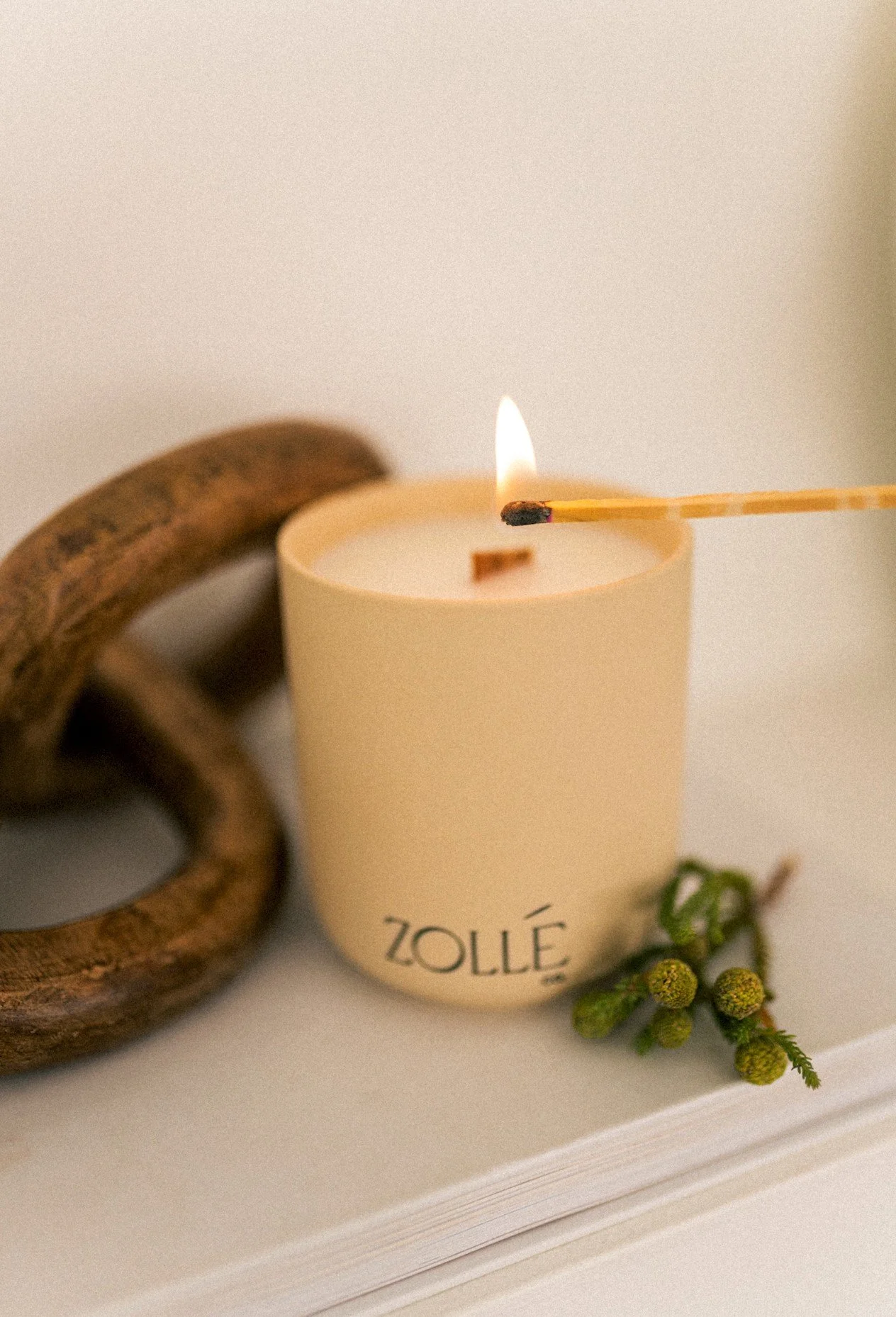 Butter-yellow Zollé candle being lit with a long match beside a carved wooden chain, candle product photography by A. Wood