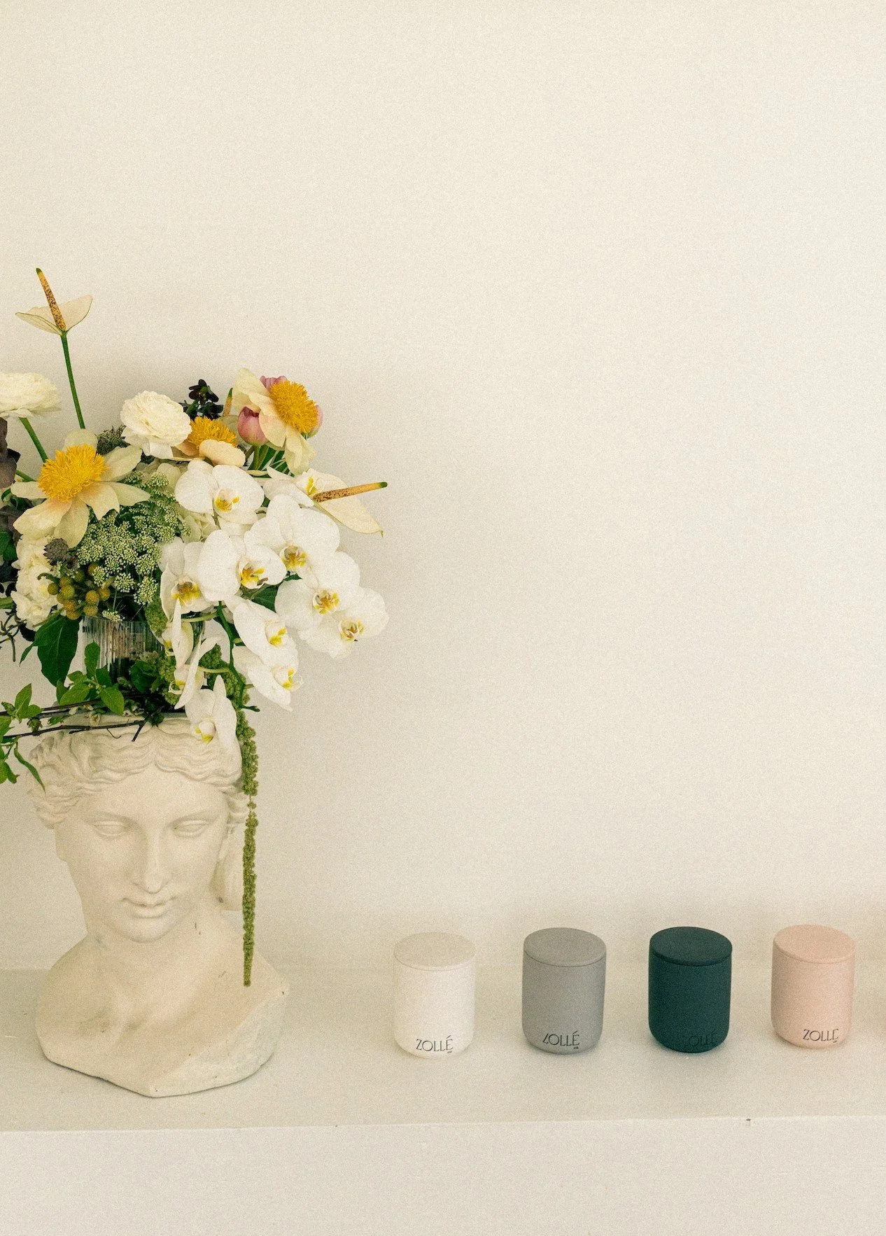 Wildflowers spilling from a white classical bust beside a row of small Zollé candles on a white shelf — candle product photography Montréal by A. Wood