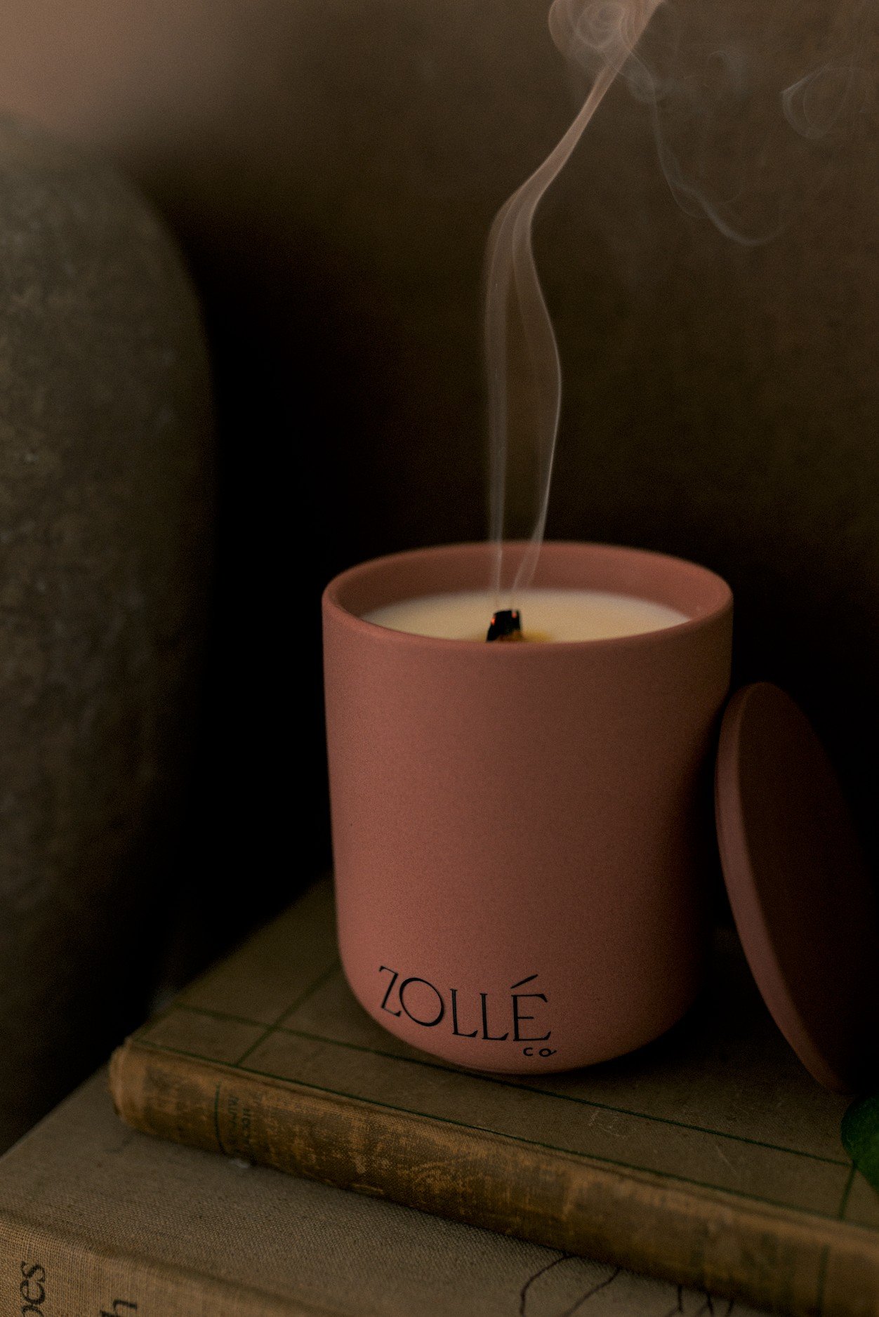 Rose-coloured Zollé candle with a curl of smoke rising from the wick, lid propped against it on an old book — moody lifestyle product photography