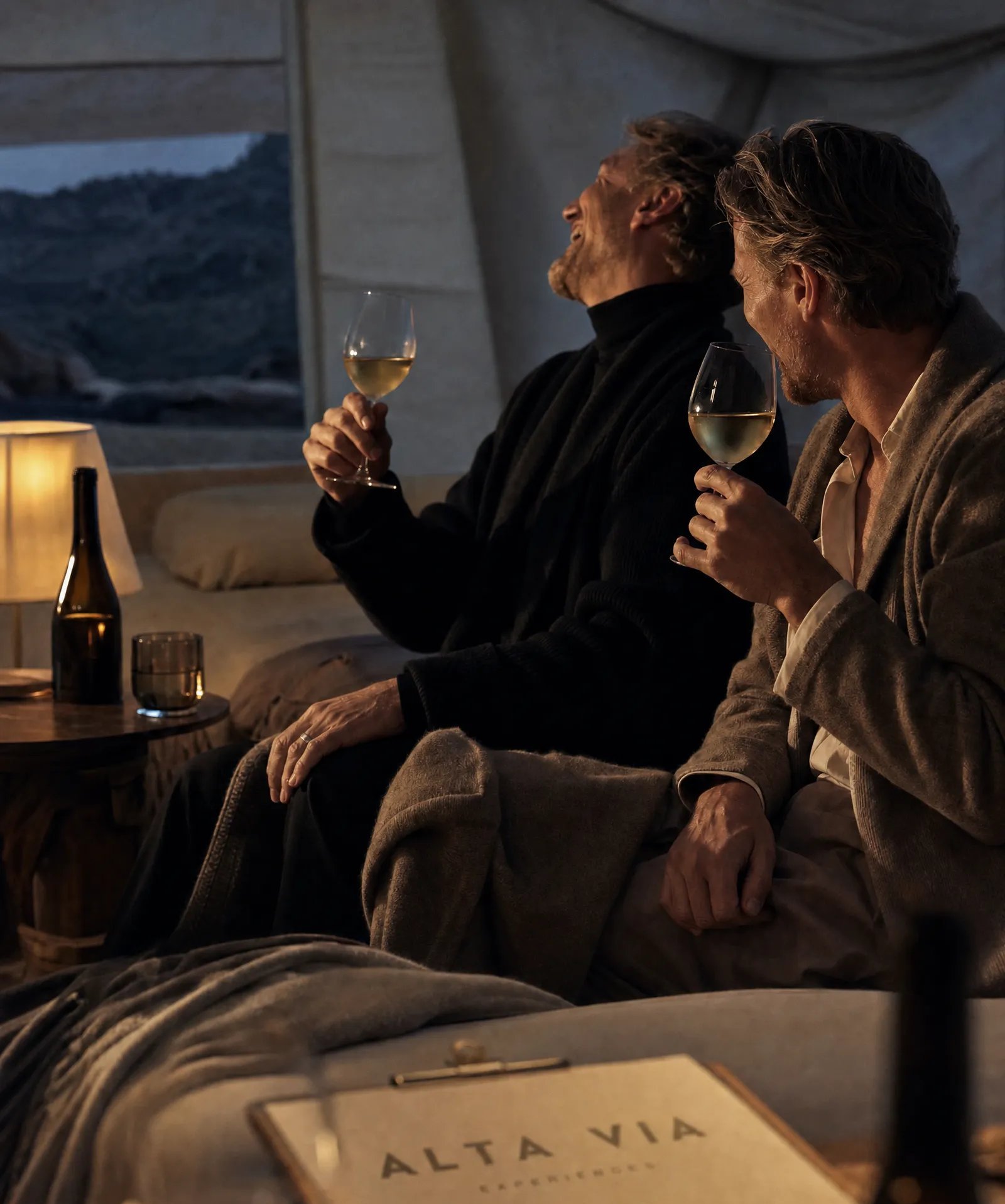 Two men laughing over wine in a canvas tent at dusk