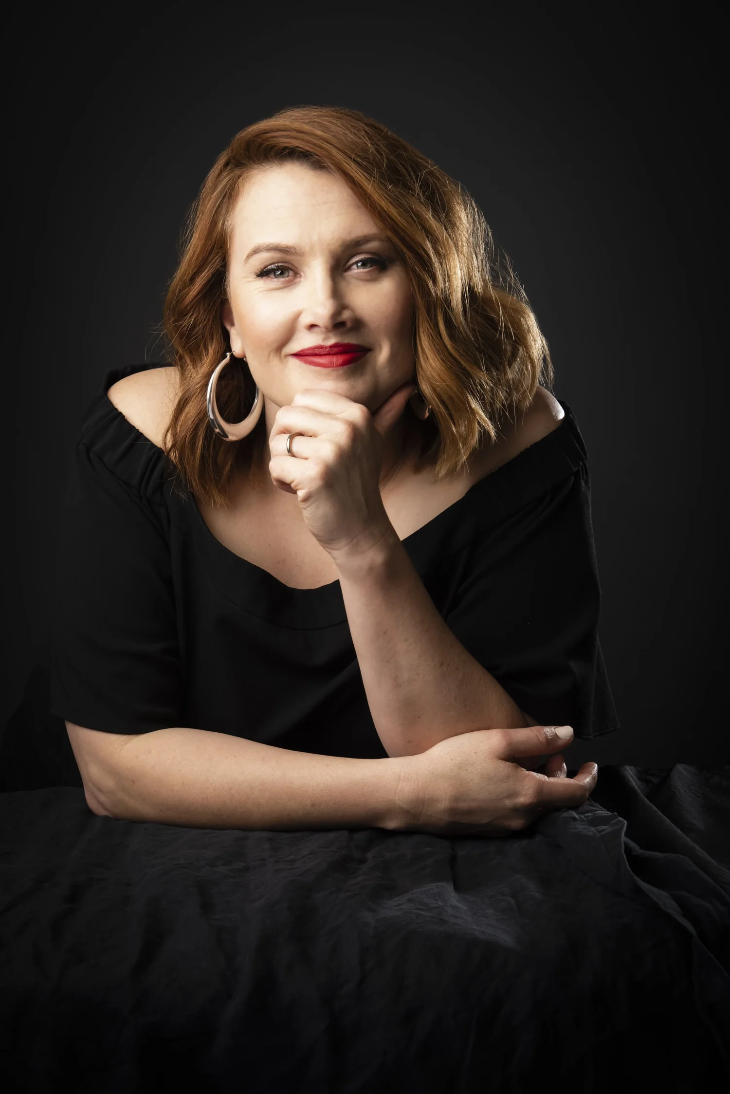 Clare Bowditch in a black top against a dark background, chin resting on her hand