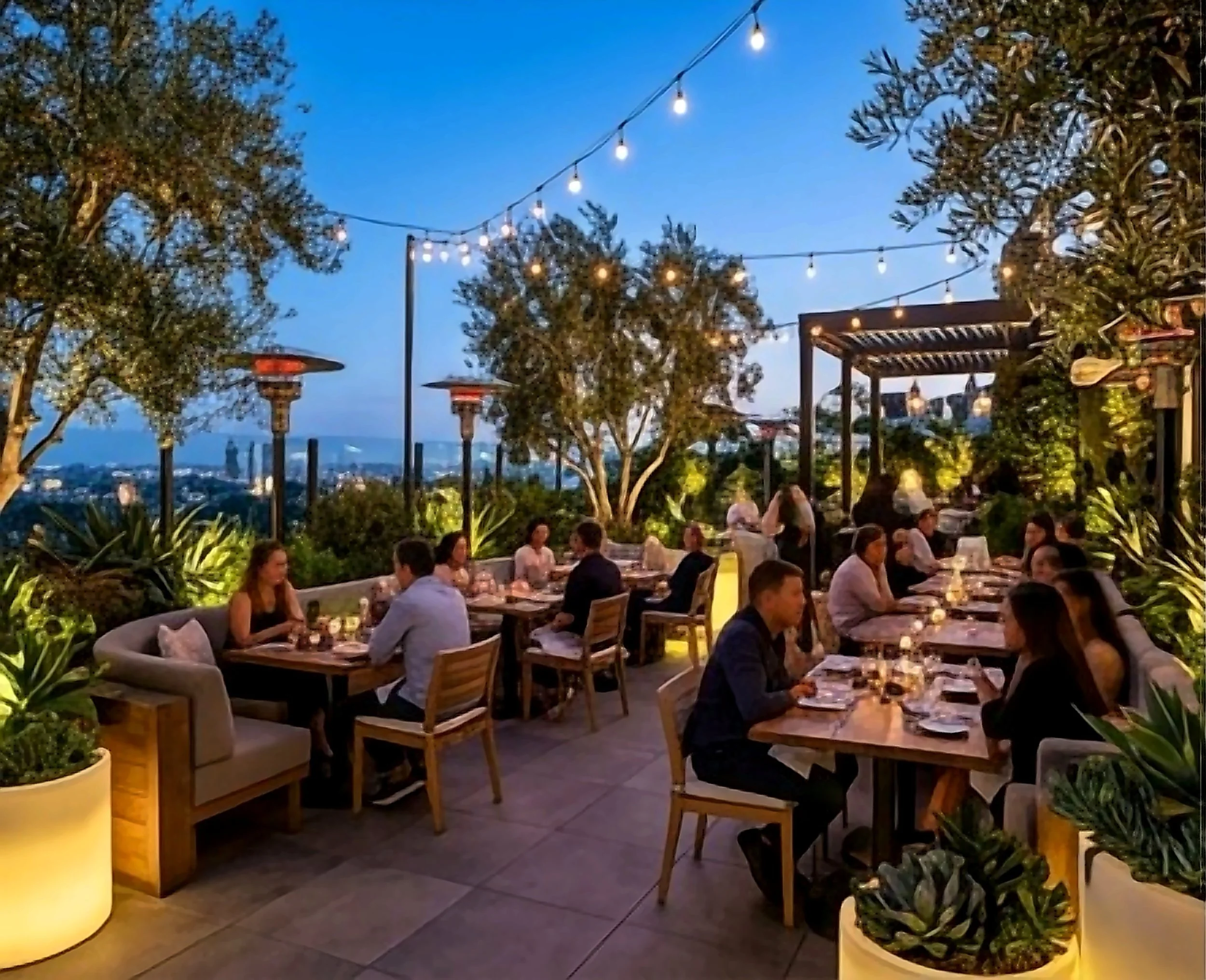 Rooftop terrace at dusk with string lights, olive trees, and city skyline view