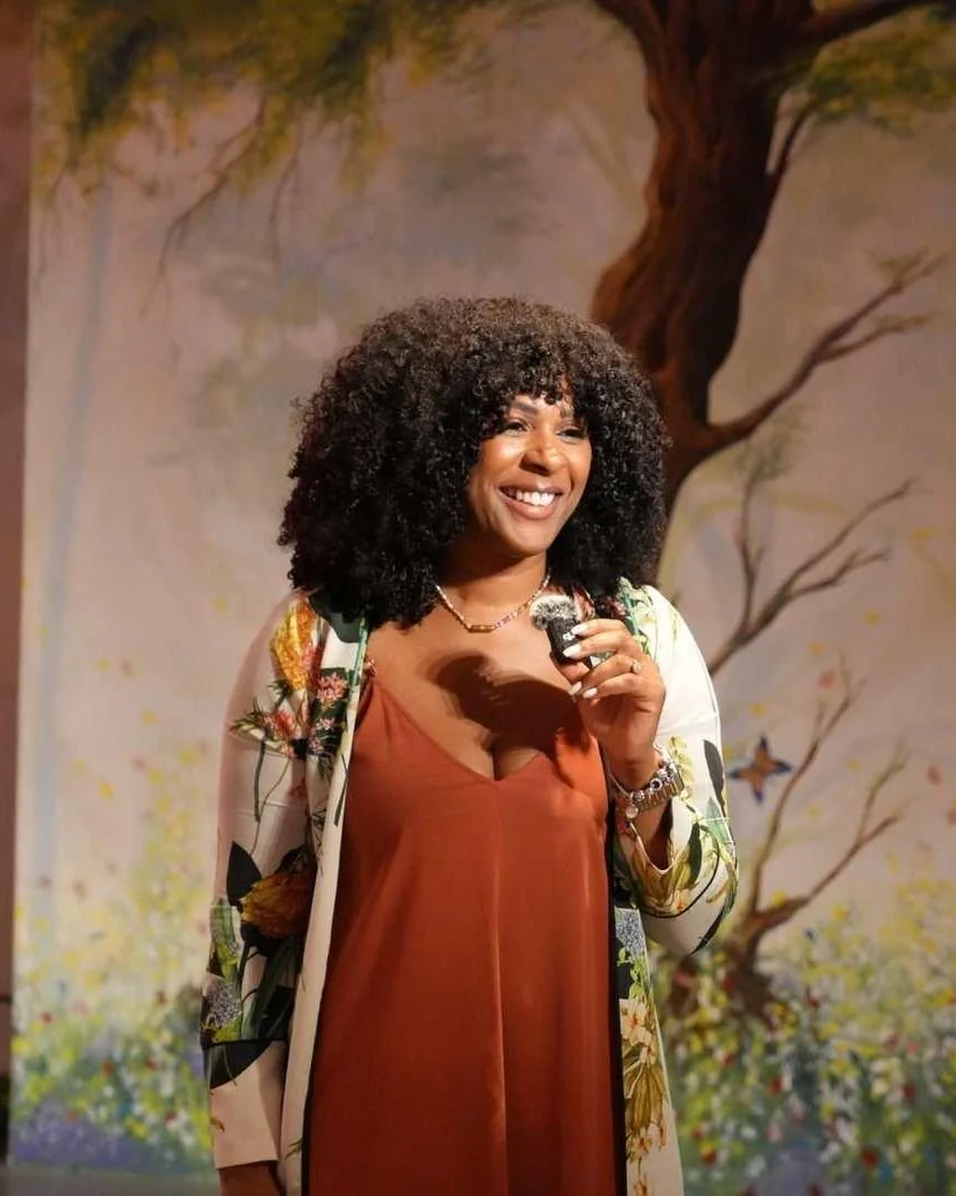 Nicole Bonnah smiling with a microphone in front of a painted tree mural at Voices of Light