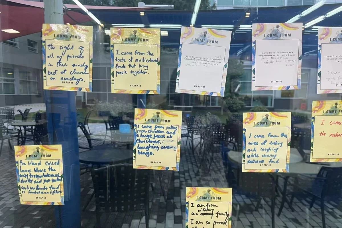 Handwritten I Come From cards displayed on a glass wall, with lines about family meals, Christmas feasts and an island home
