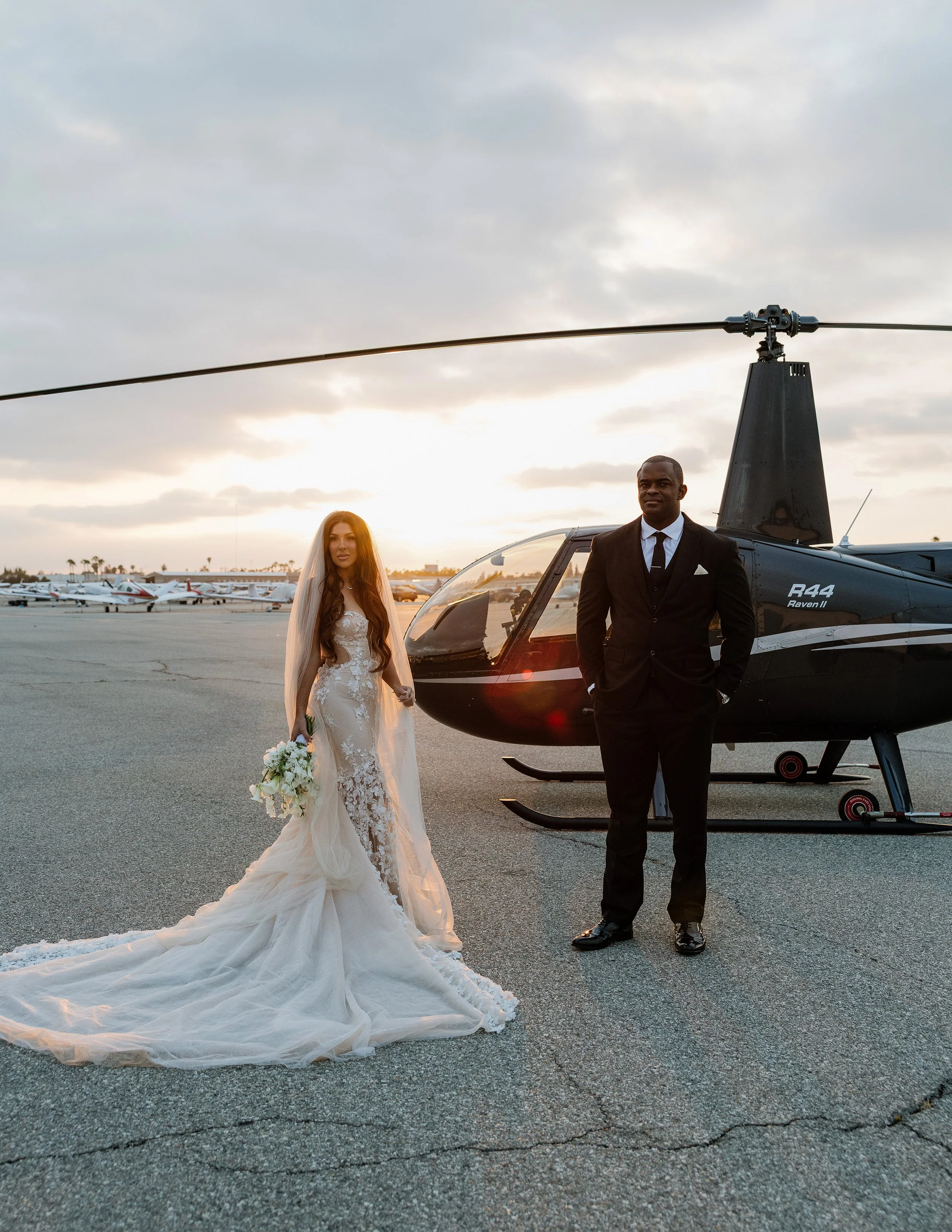 Luxury wedding portrait of a bride and groom standing beside a helicopter on an airport runway at sunset.