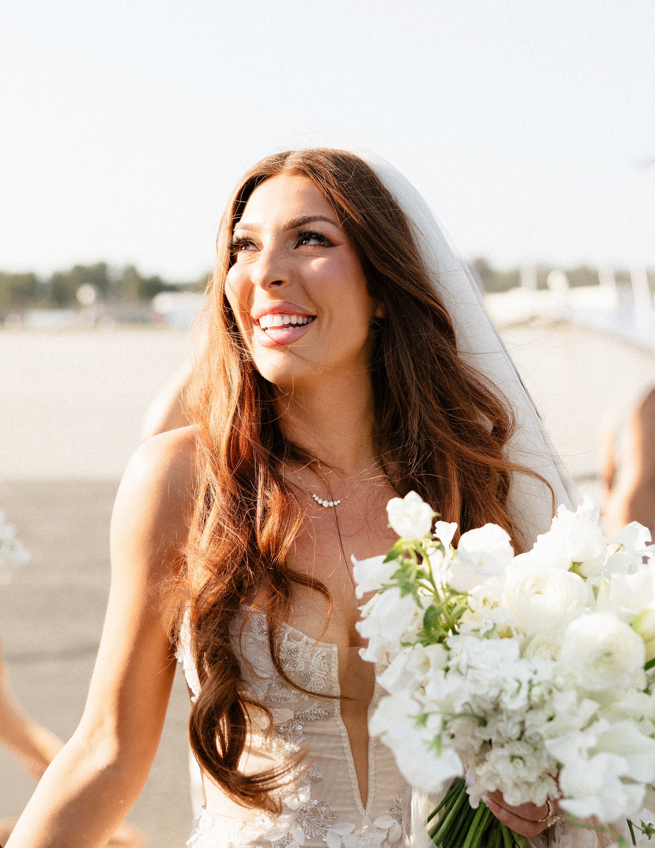 Warm outdoor bridal portrait of a smiling bride holding a white bouquet in natural sunlight.