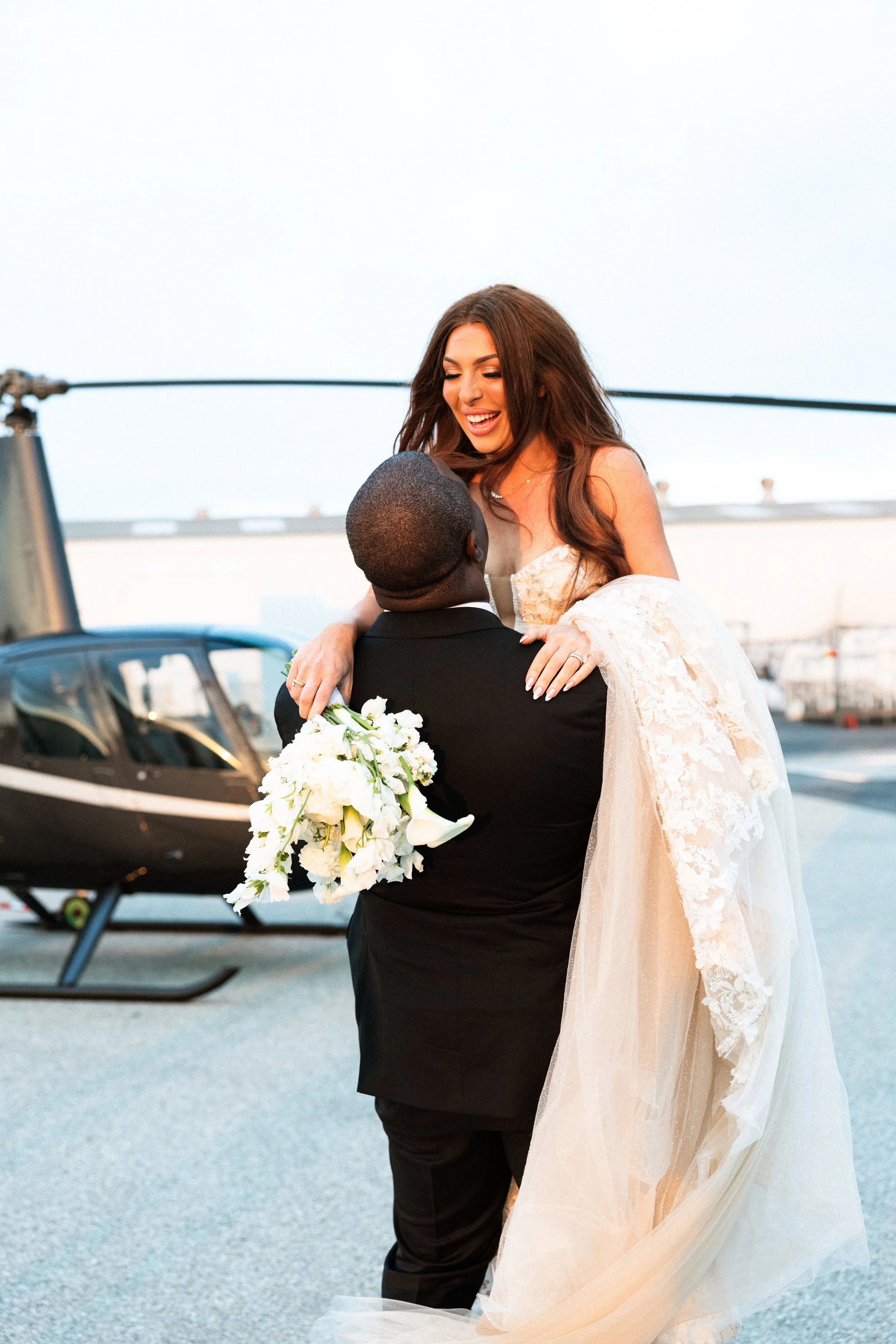 Candid luxury wedding portrait of a groom lifting his bride beside a helicopter on an airport tarmac.