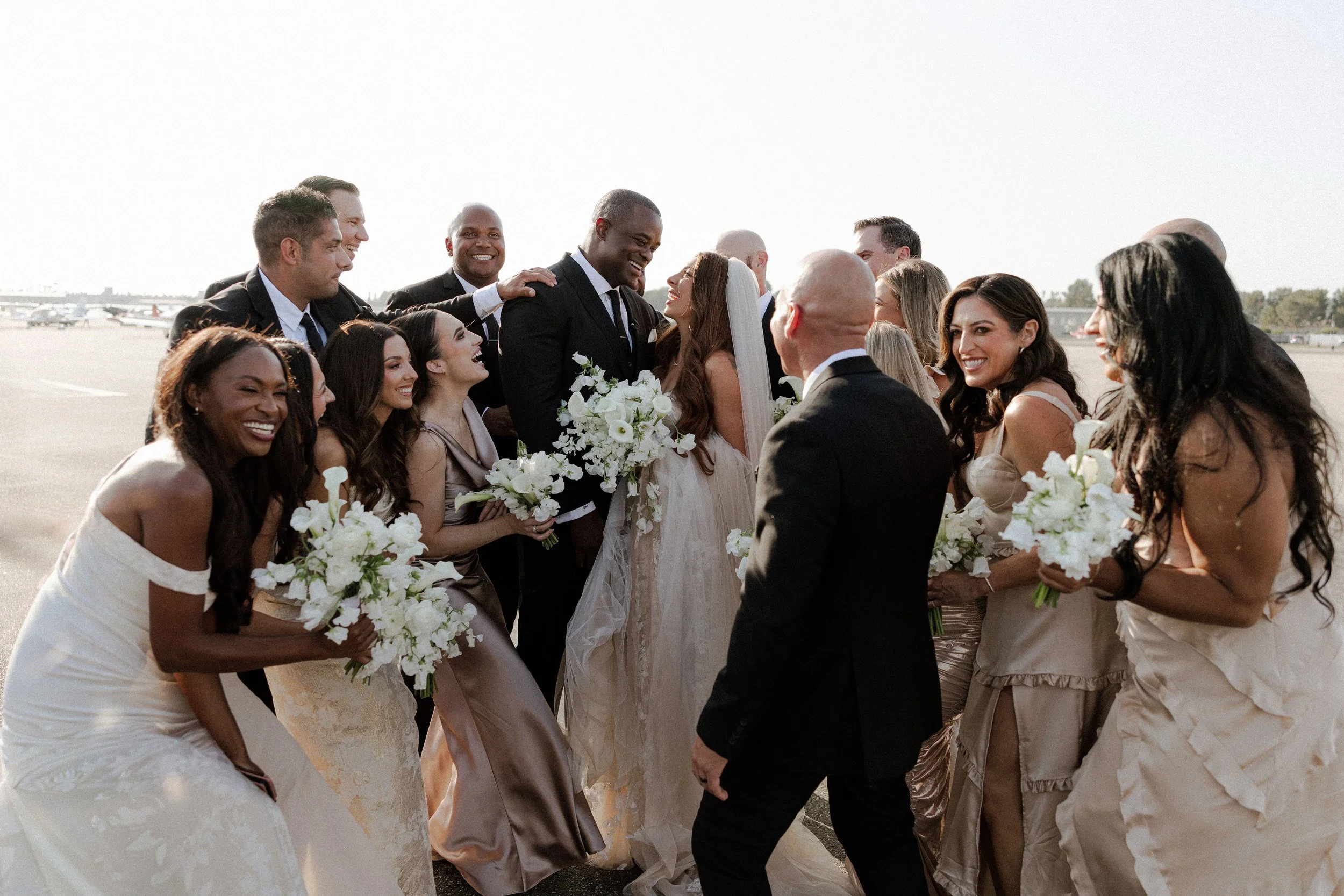 Candid outdoor wedding party photo of a bride and groom laughing with bridesmaids and groomsmen in natural sunlight.