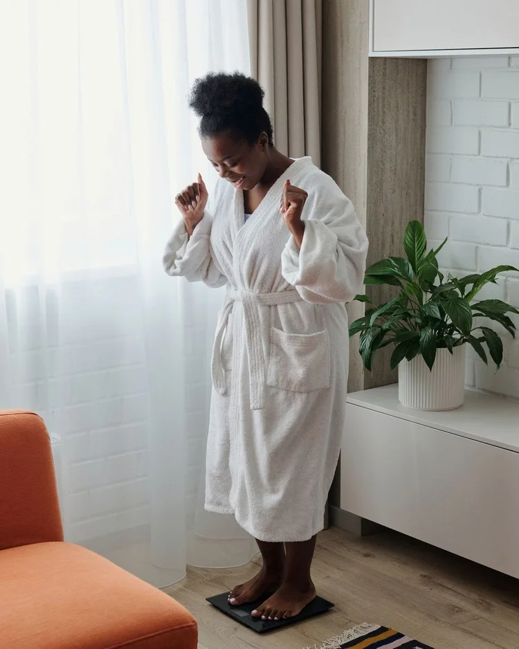 A smiling woman in a bathrobe celebrating as she steps on a bathroom scale at home