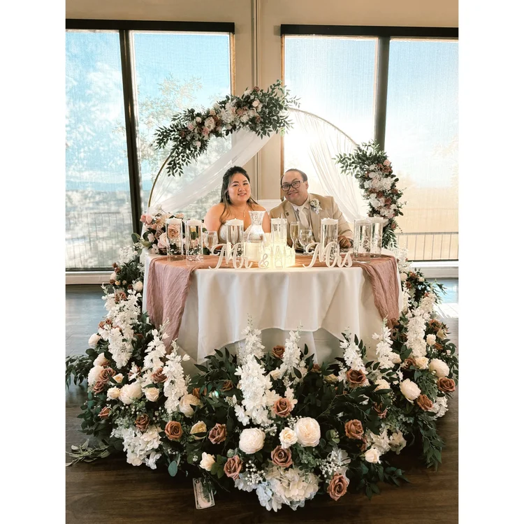 Newlyweds seated at a sweetheart table surrounded by white and blush floral arrangements beneath a draped arch