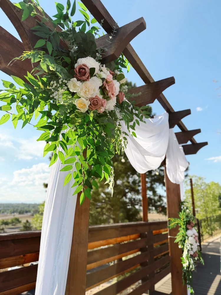 Wooden pergola ceremony arch styled with blush roses, white blooms, greenery, and flowing white draping