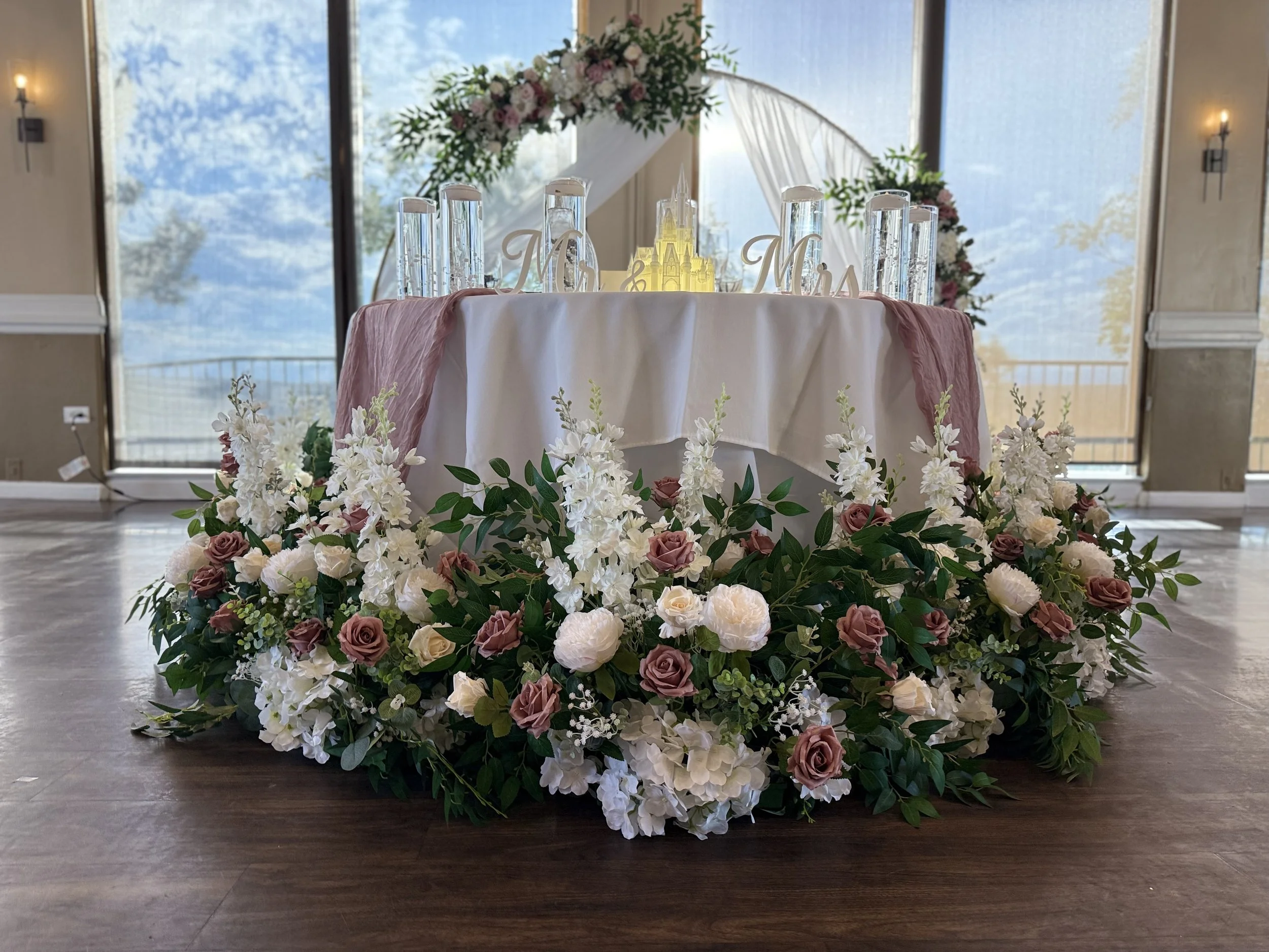 Long low sweetheart table garland of dusty rose and cream roses, white stock, and greenery beneath blush draping