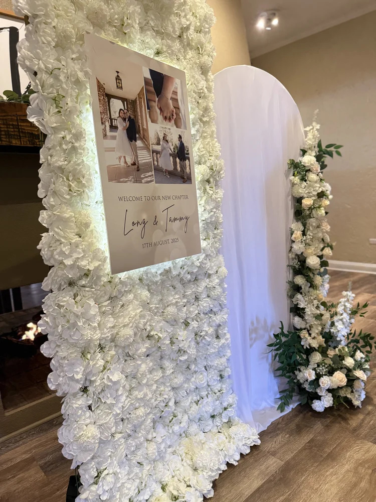Wedding welcome sign framed by a white floral wall beside a draped arch with white floral arrangements