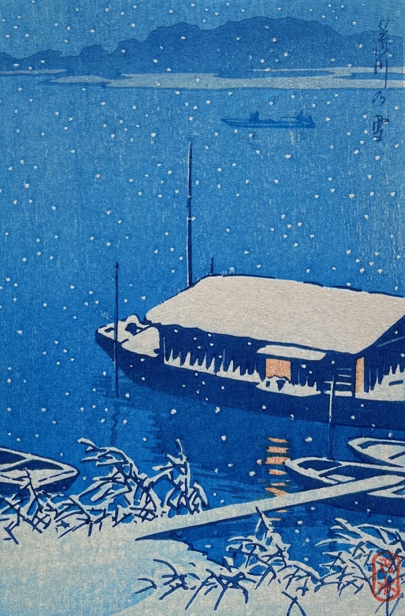 Arakawa River in Snow by Kawase Hasui: falling white snow against blue river and sky, a covered boat with a snowy roof and two warm windows, orange reflections, small boats and snow-covered foreground branches.