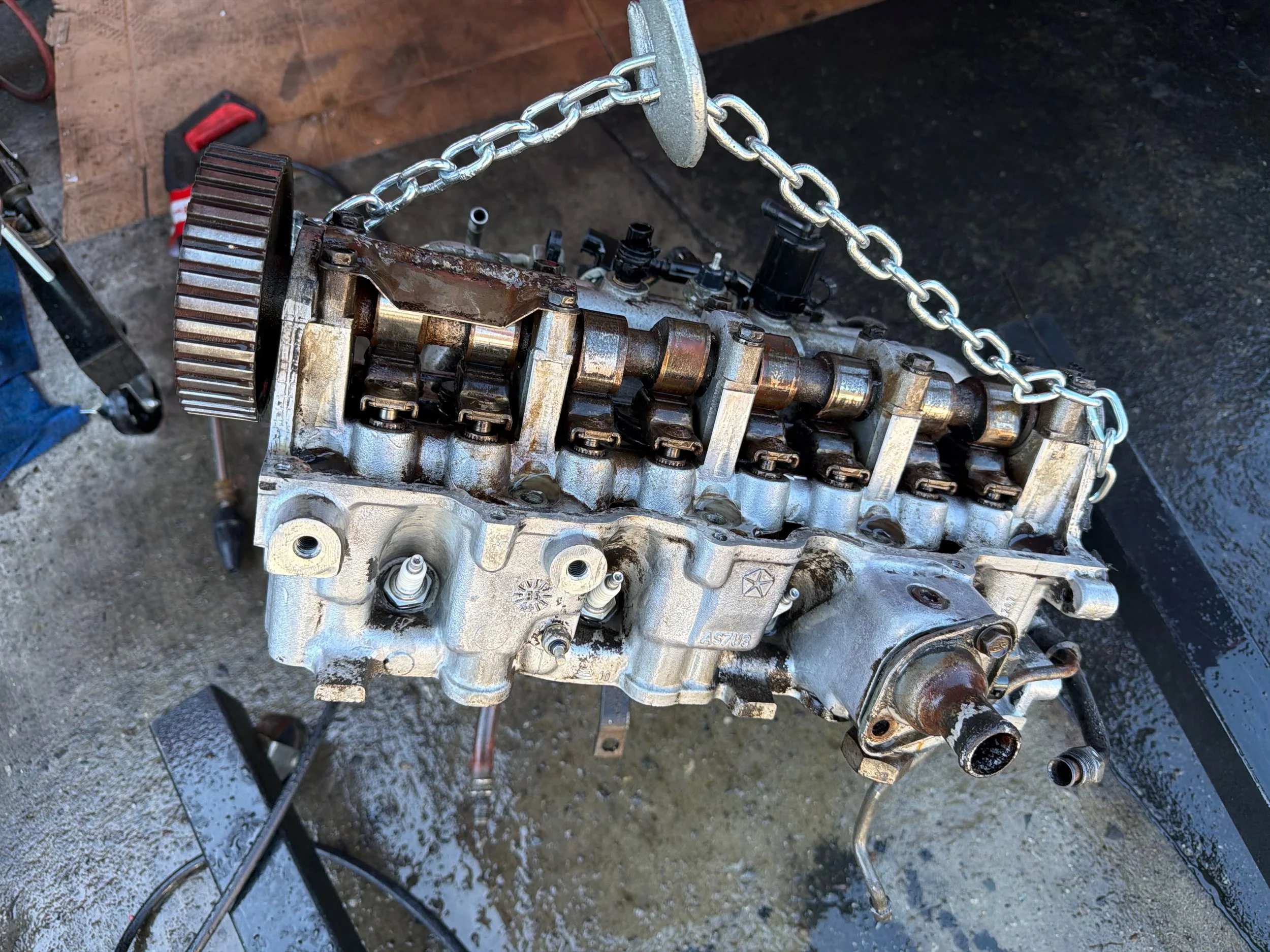 The cylinder head off the 1985 Chrysler Town and Country wagon, hanging from an engine hoist by chains, showing the camshaft and rocker arms