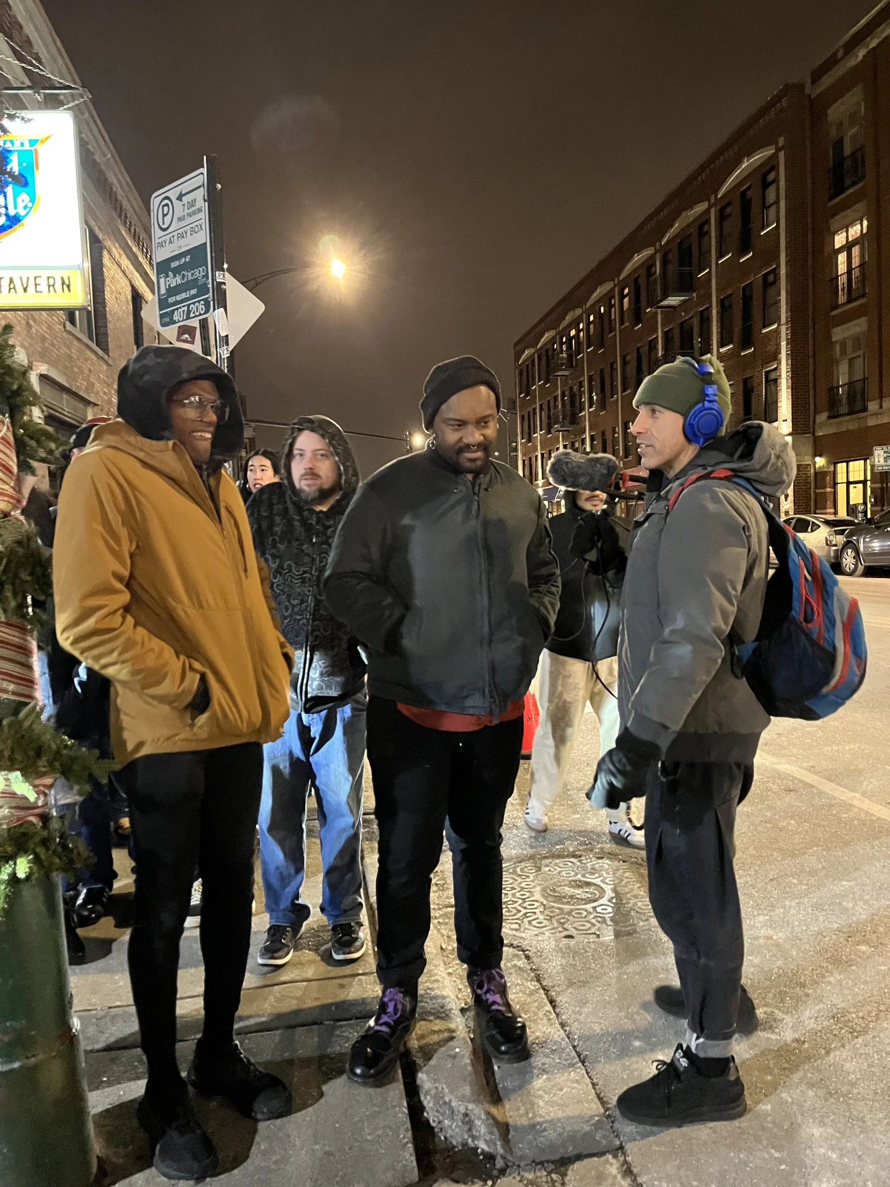 Ramtin Arablouei wearing blue headphones and interviewing a group on a city sidewalk at night.