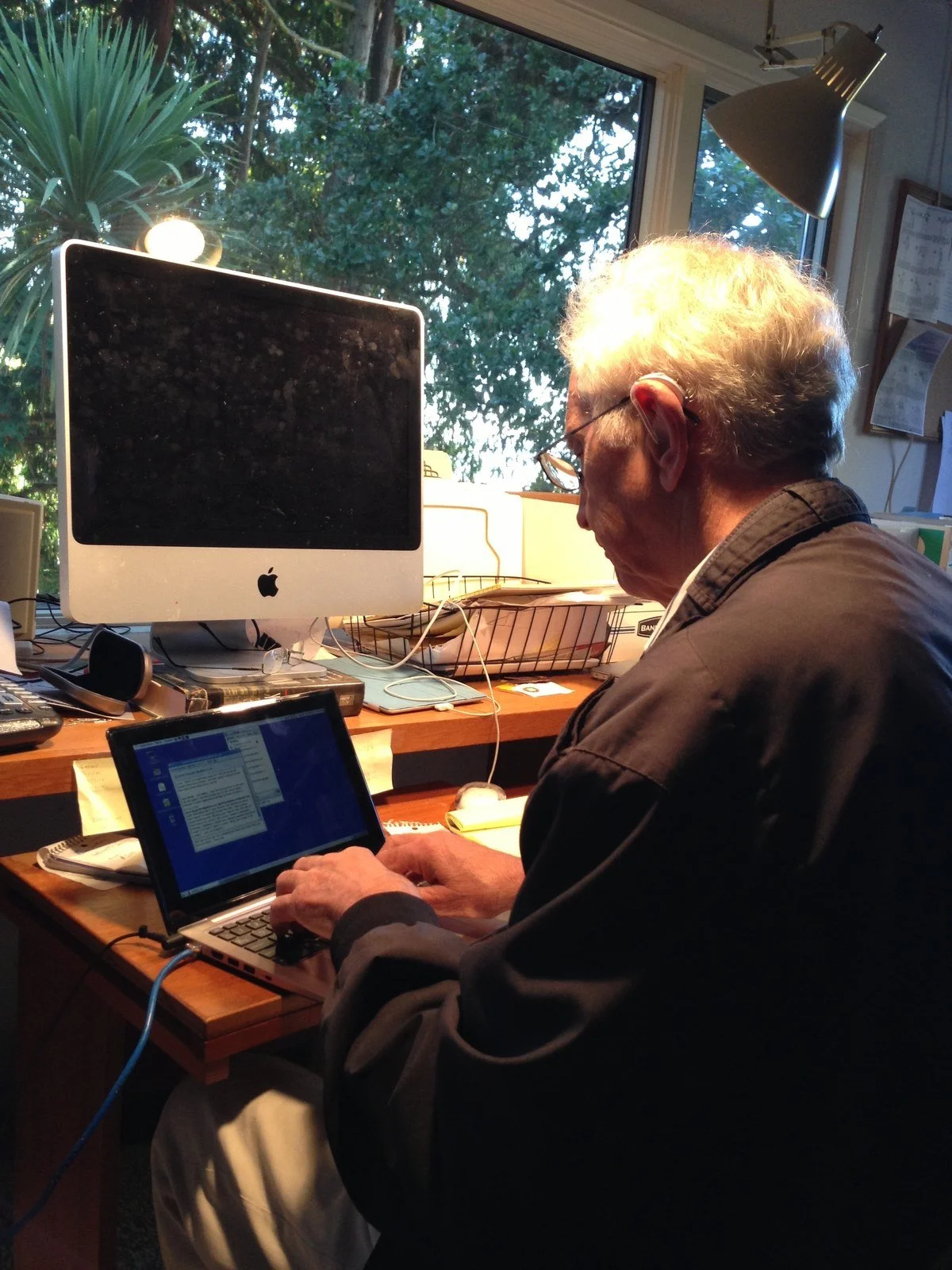 Daniel Ellsberg seated at a desk, typing on a small laptop beside a large monitor, with windows behind the desk.