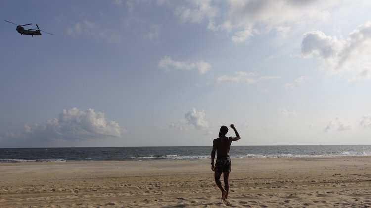 Rashad Houston stands barefoot on the beach with one fist raised as a twin-rotor helicopter passes overhead.