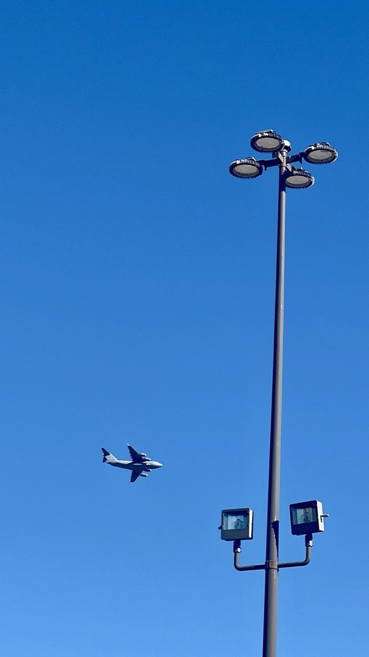 An aircraft passing beside a tall lighting tower against a blue sky.