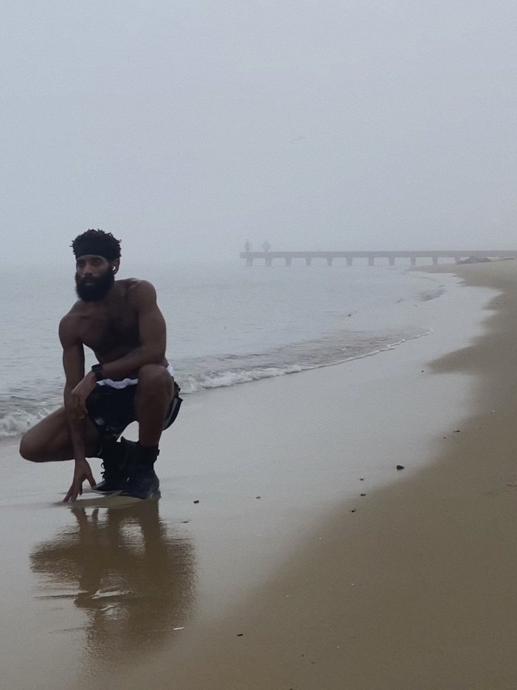 Rashad Houston crouches at the water’s edge on a foggy beach, with his reflection in the wet sand.