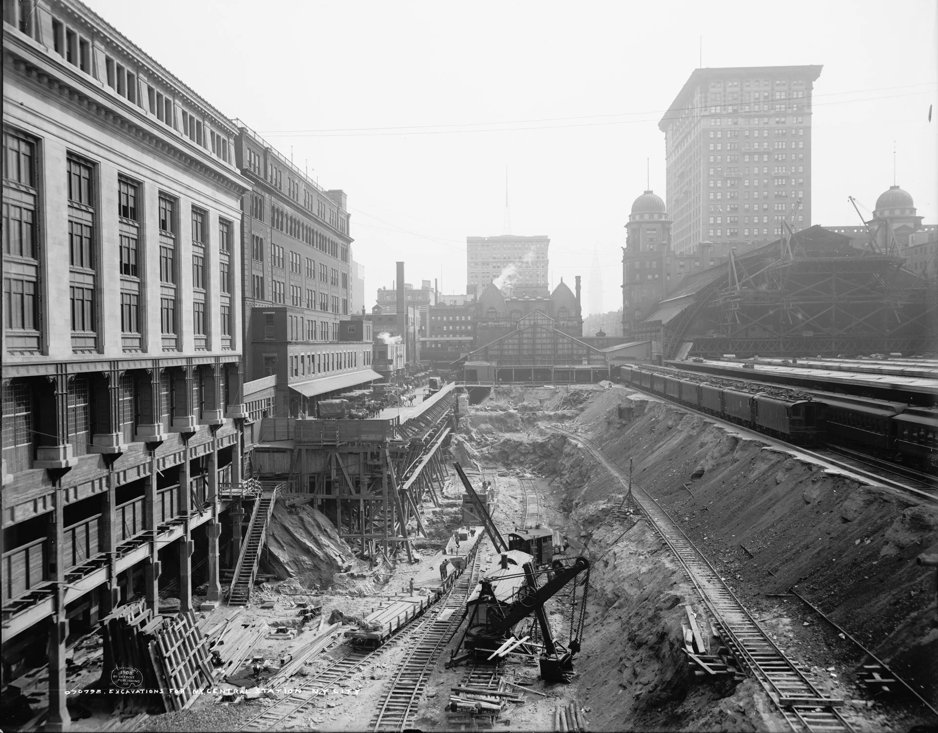 Workers and rail-mounted excavation machinery in a deep construction cutting beside the old Grand Central station and tracks, 1908.