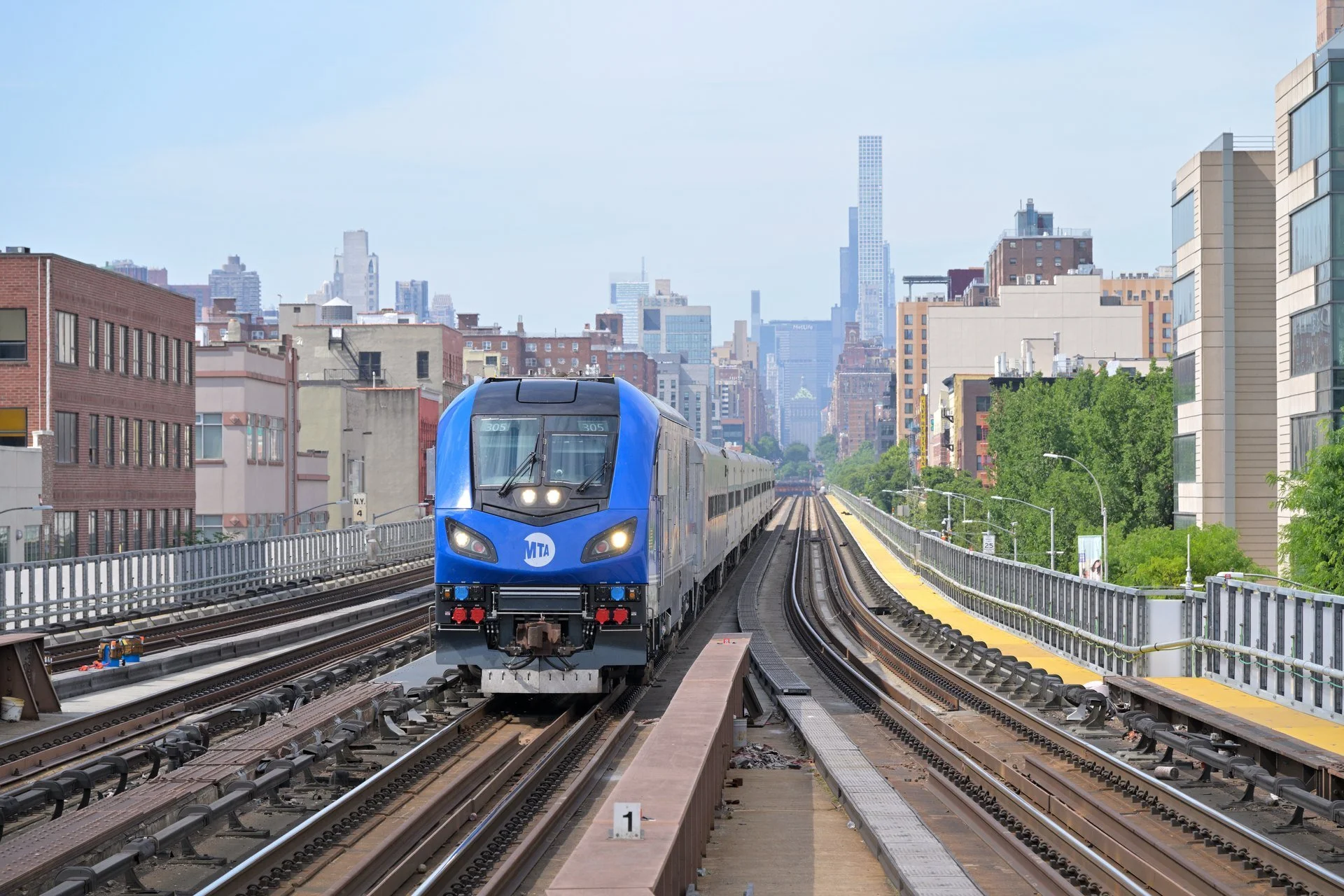 A blue Metro-North locomotive approaches Harlem–125th Street along elevated tracks, with Manhattan buildings beyond.