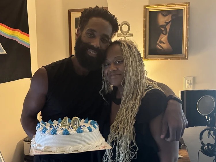 Rashad and a woman with long locs sharing an embrace beside a birthday cake.