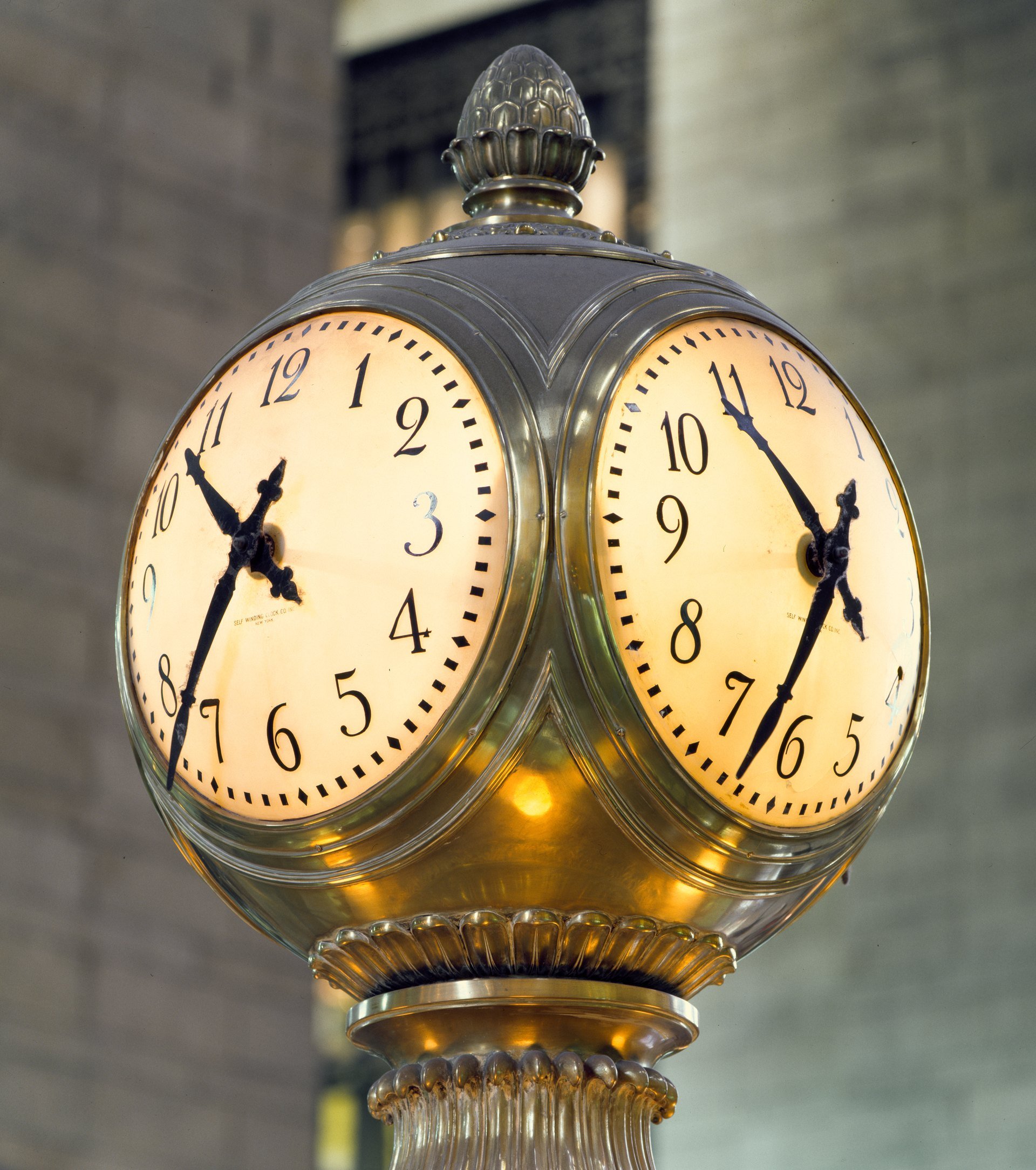 Close view of two illuminated glass faces of the brass clock, with an acorn finial above.