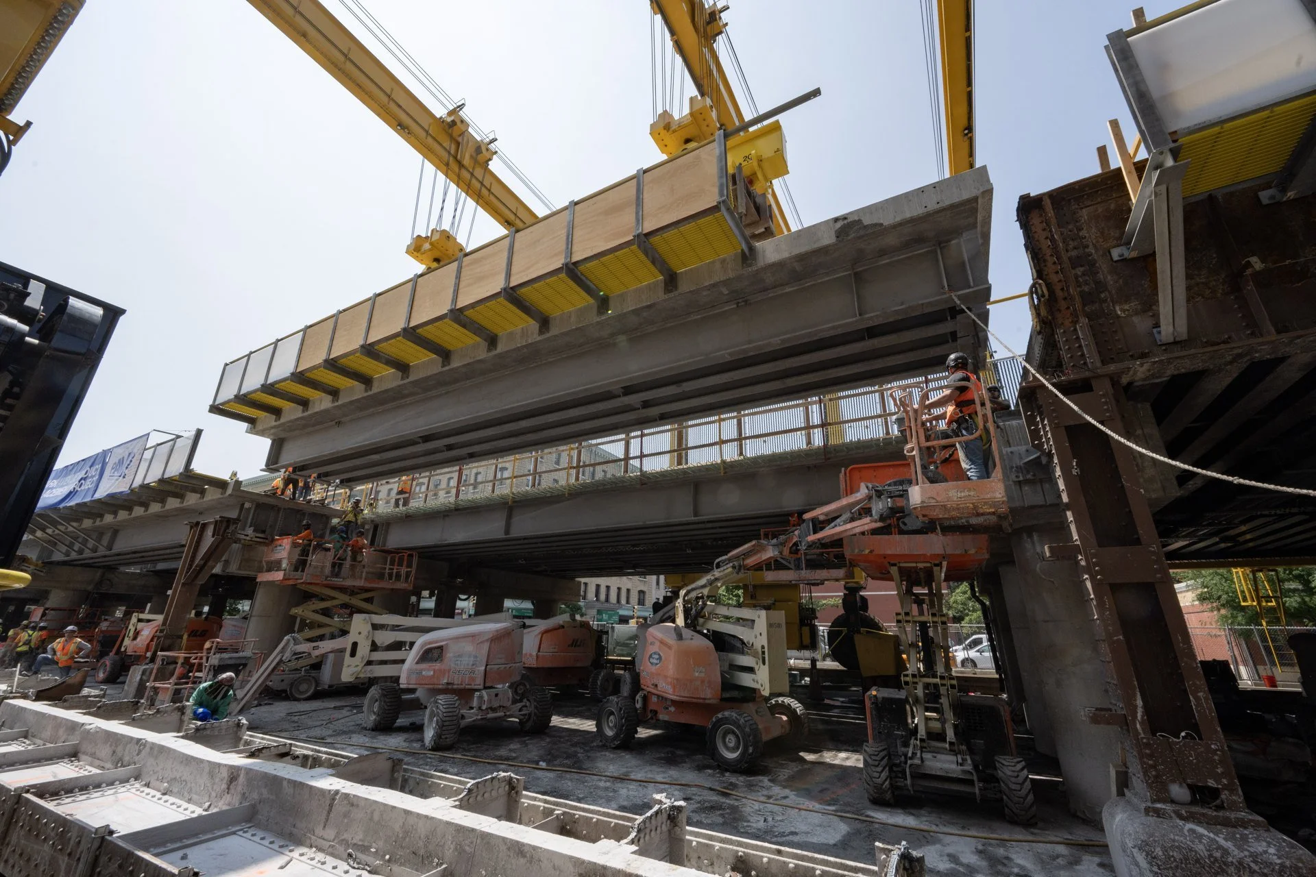 Workers in elevated platforms guide a large prefabricated bridge section suspended from yellow gantry equipment in East Harlem.
