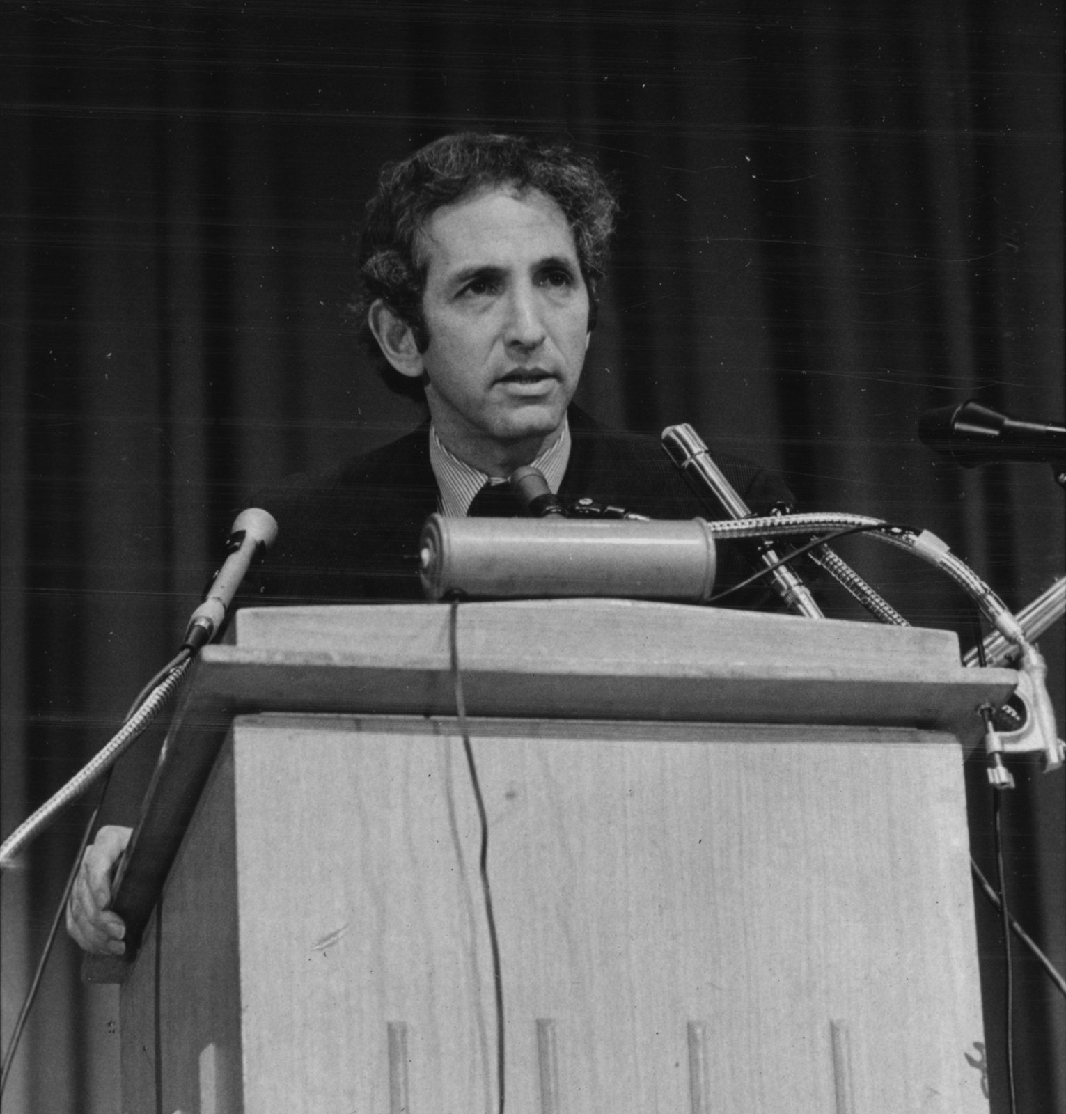 Daniel Ellsberg standing behind a lectern with several microphones at a press conference.