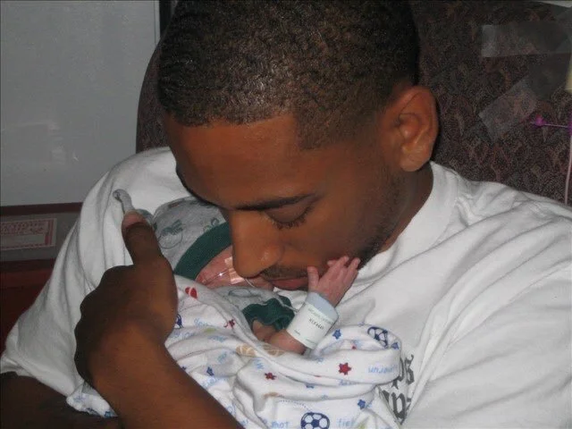 Rashad holding his older son at birth in a patterned hospital blanket.