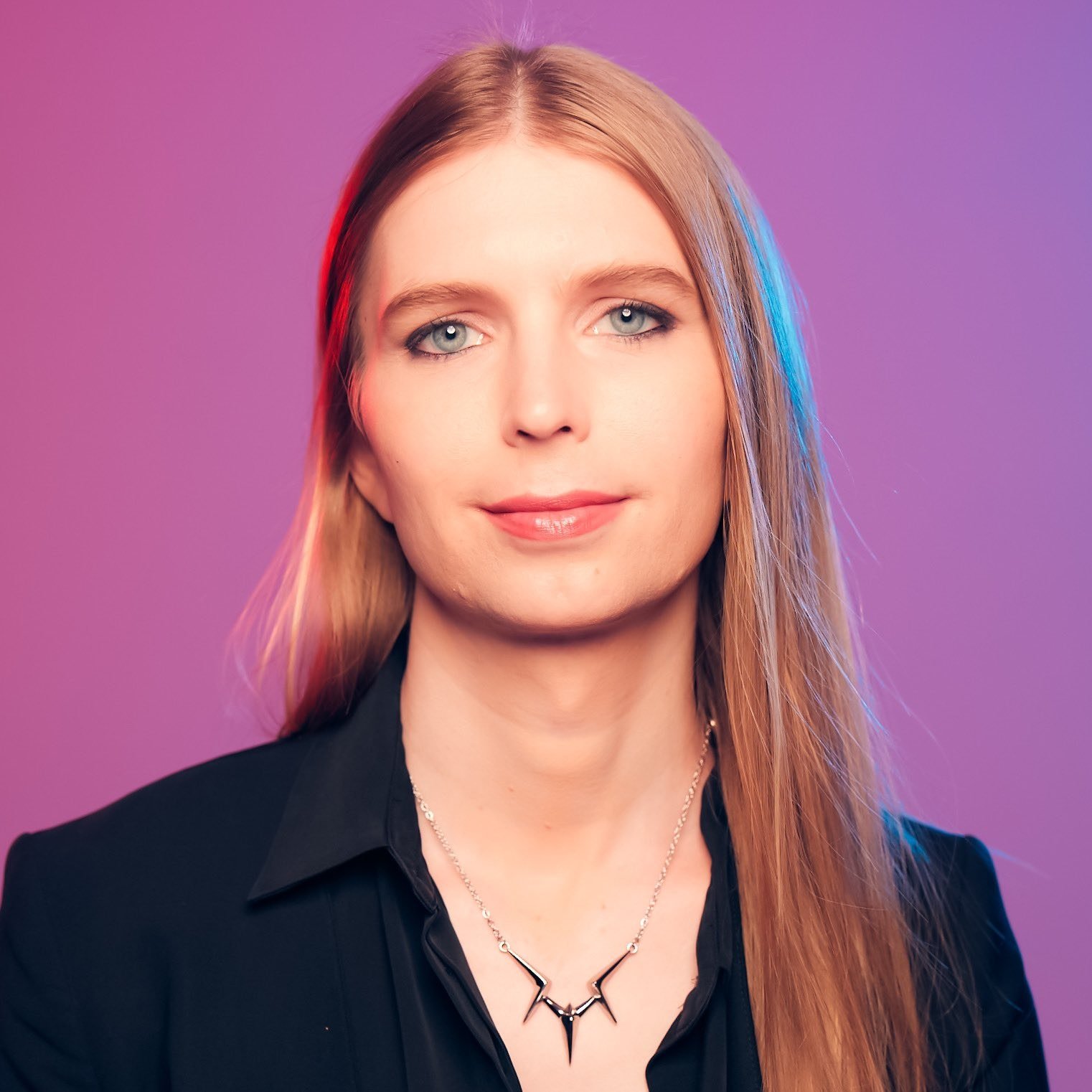 Chelsea Manning with long reddish hair, wearing a black collared shirt against a purple background.
