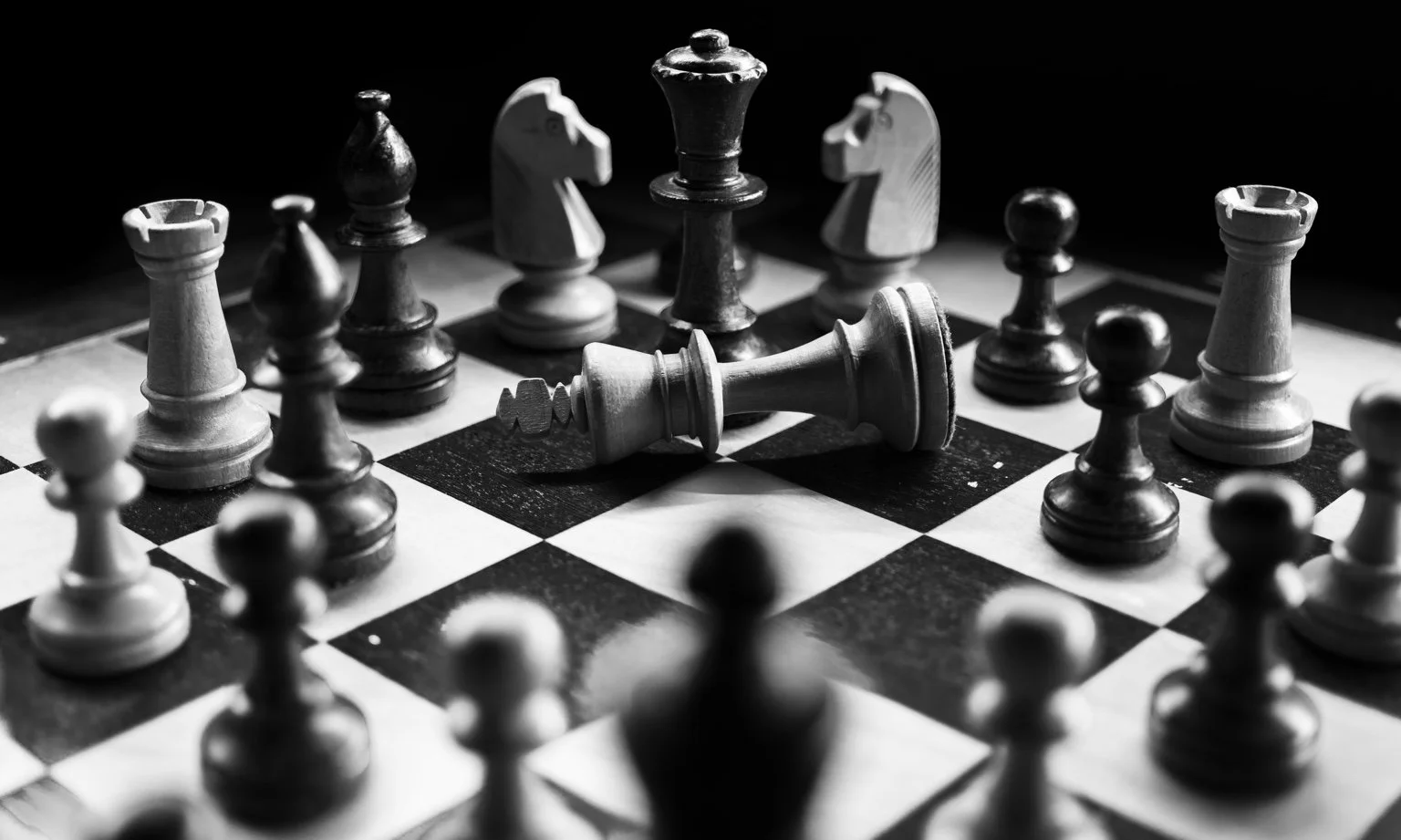 A fallen white king lies on a chessboard surrounded by upright black and white pieces, photographed in black and white.