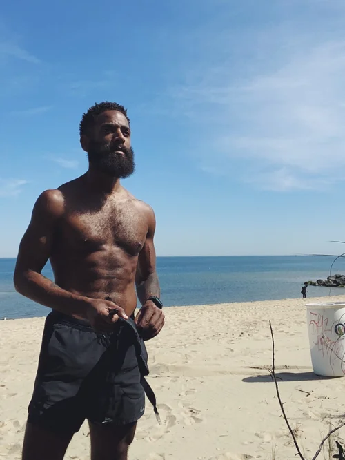 Rashad Houston standing on the sandy beach beneath a blue sky, with the bay behind him.