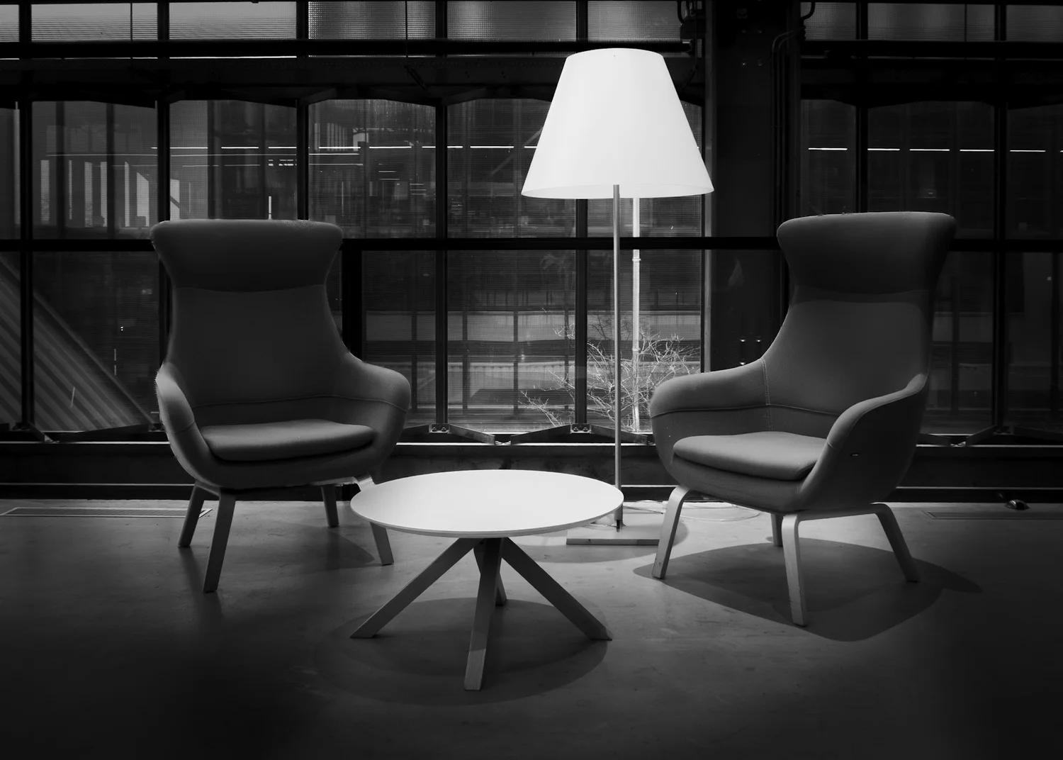 Two chairs facing each other across a small table beneath a lamp, in front of tall city windows