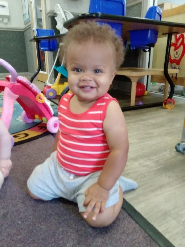 Original phone snap of a baby boy kneeling and smiling on a daycare classroom floor