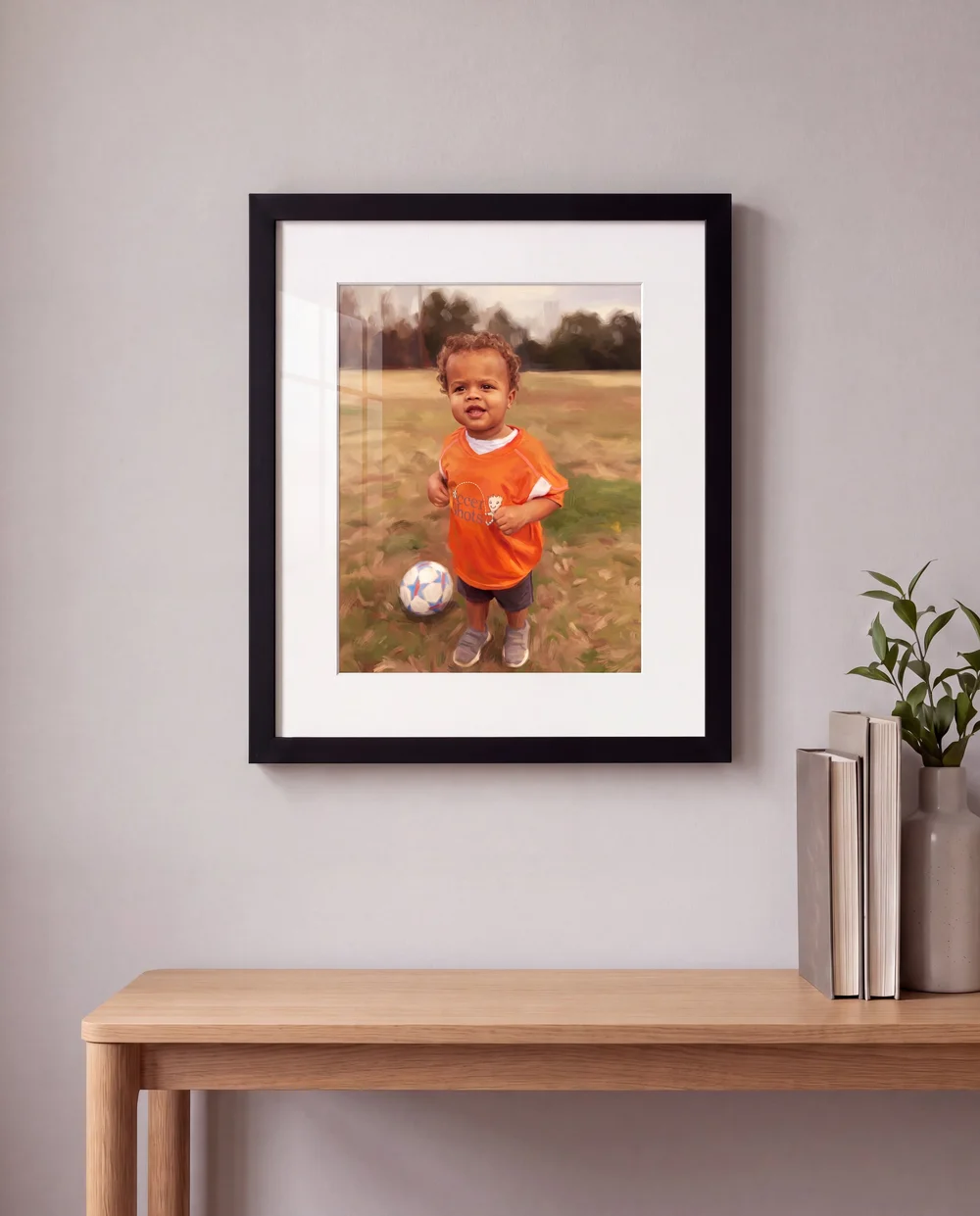 A framed illustrated portrait of a boy in an orange soccer jersey, in a black frame with a wide white mat, hanging above a console table with books and a vase